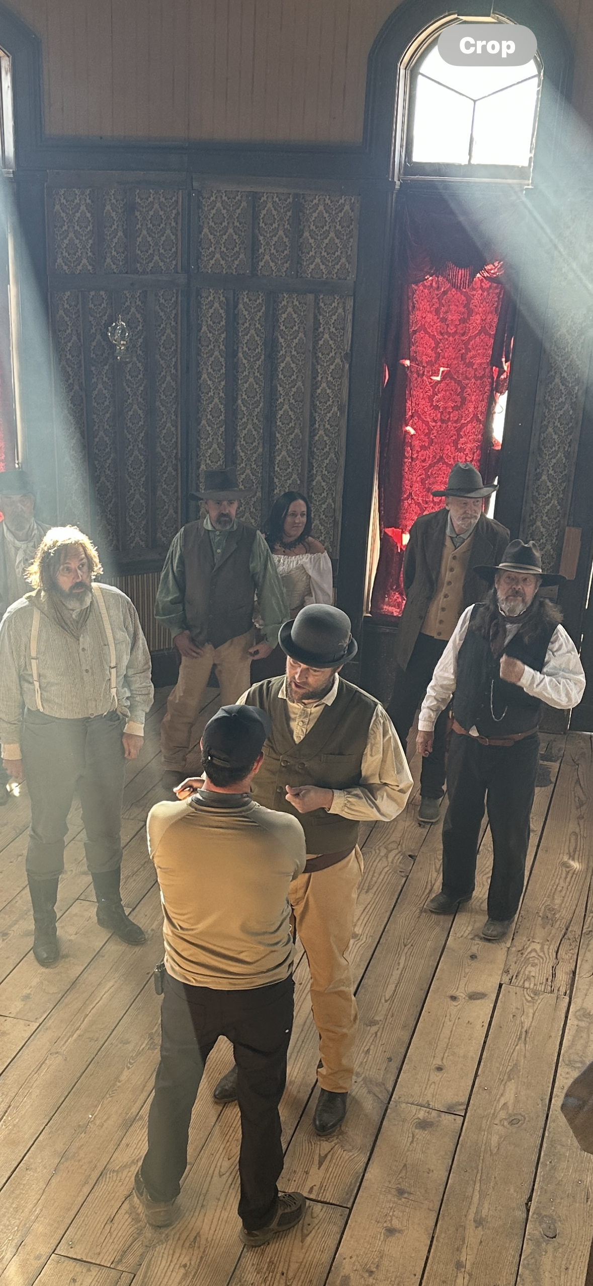 A scene from a play set in the Old West, featuring actors dressed in period costumes with hats, vests, and suspenders, standing on a wooden stage with dark, vintage-style decor and red curtains.