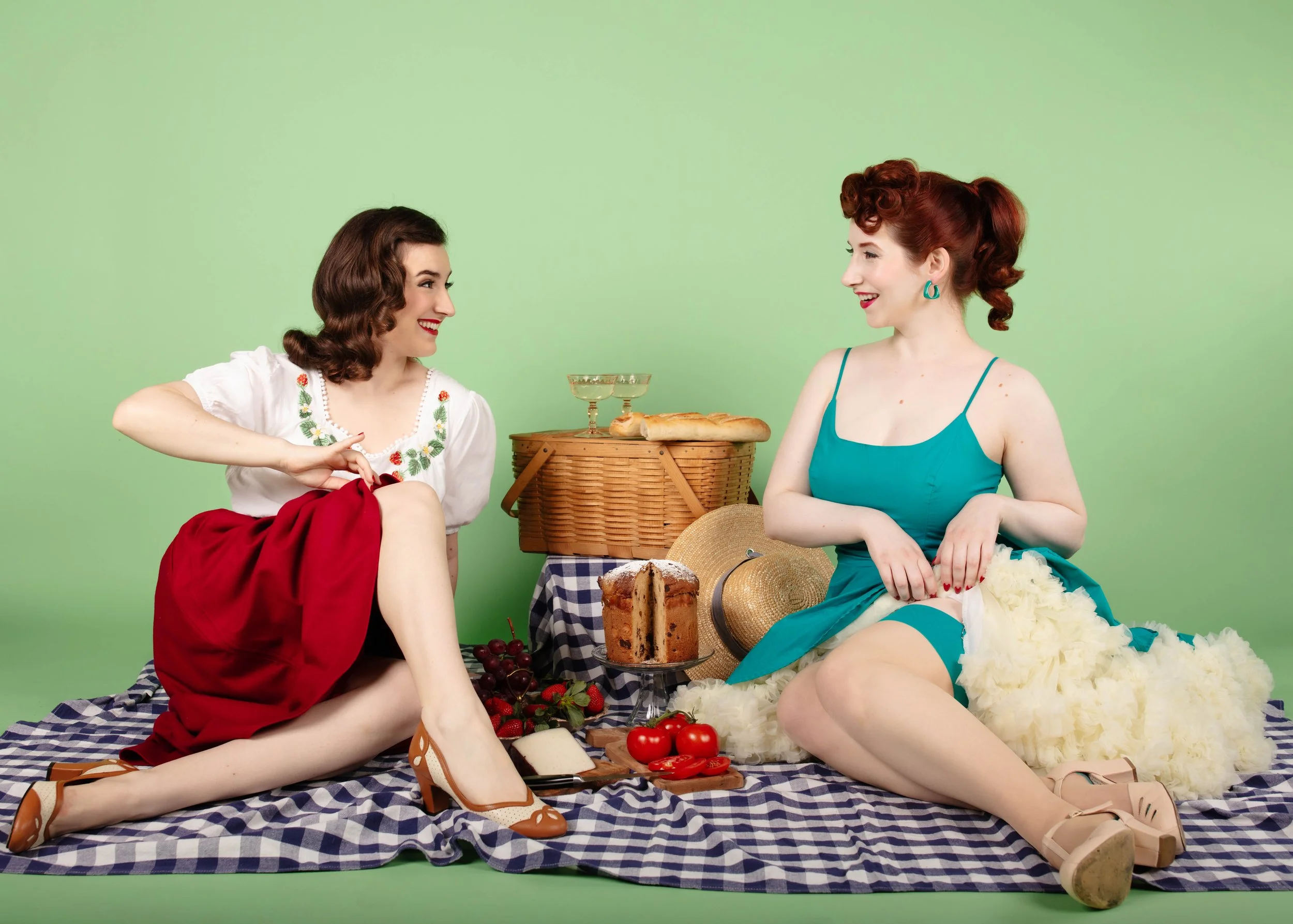 Two women having a picnic on a checkered blanket with bread, cake, fruit, and drinks, against a green background.