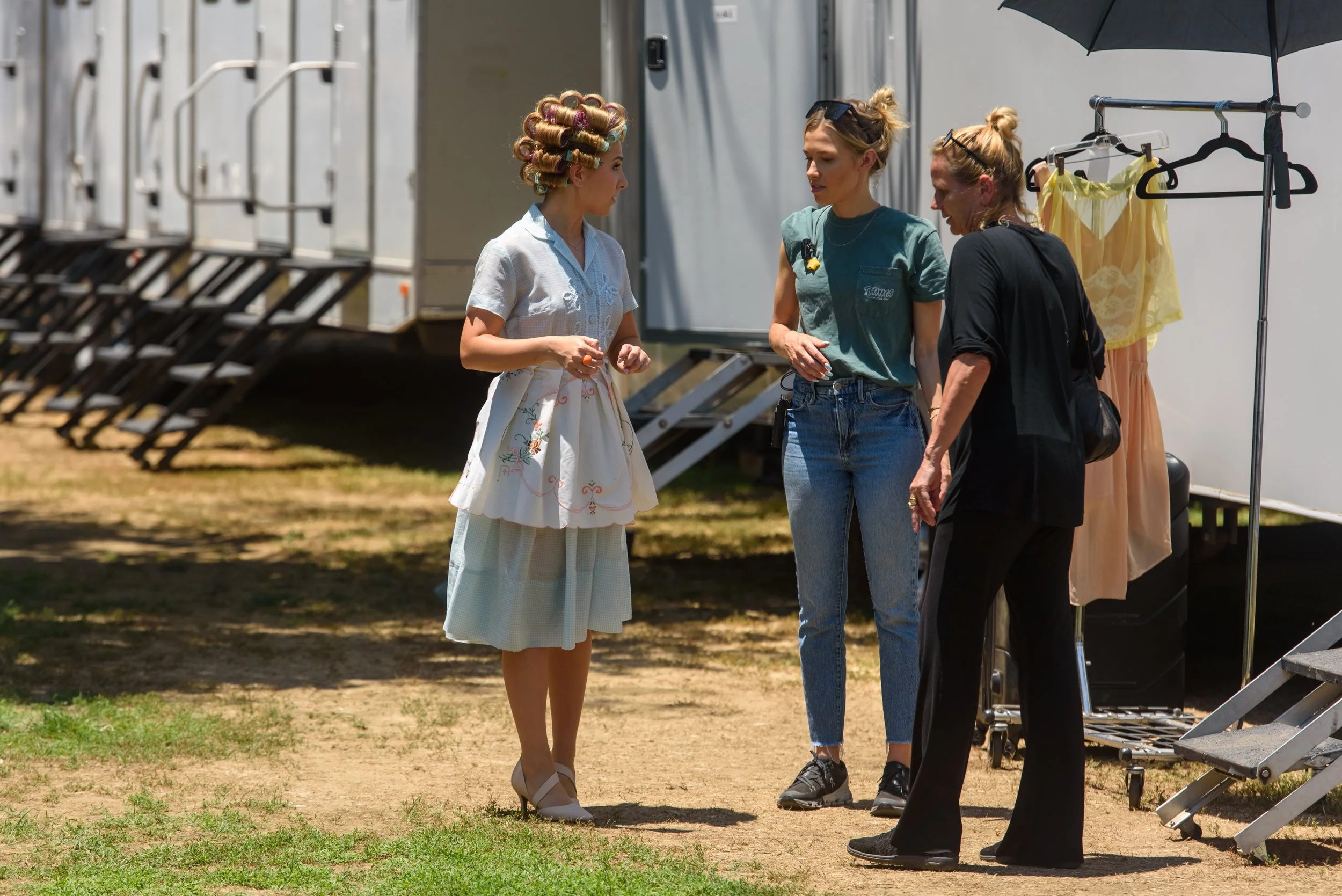 Three women engaged in conversation outside on set with trailers and clothing racks in the background.