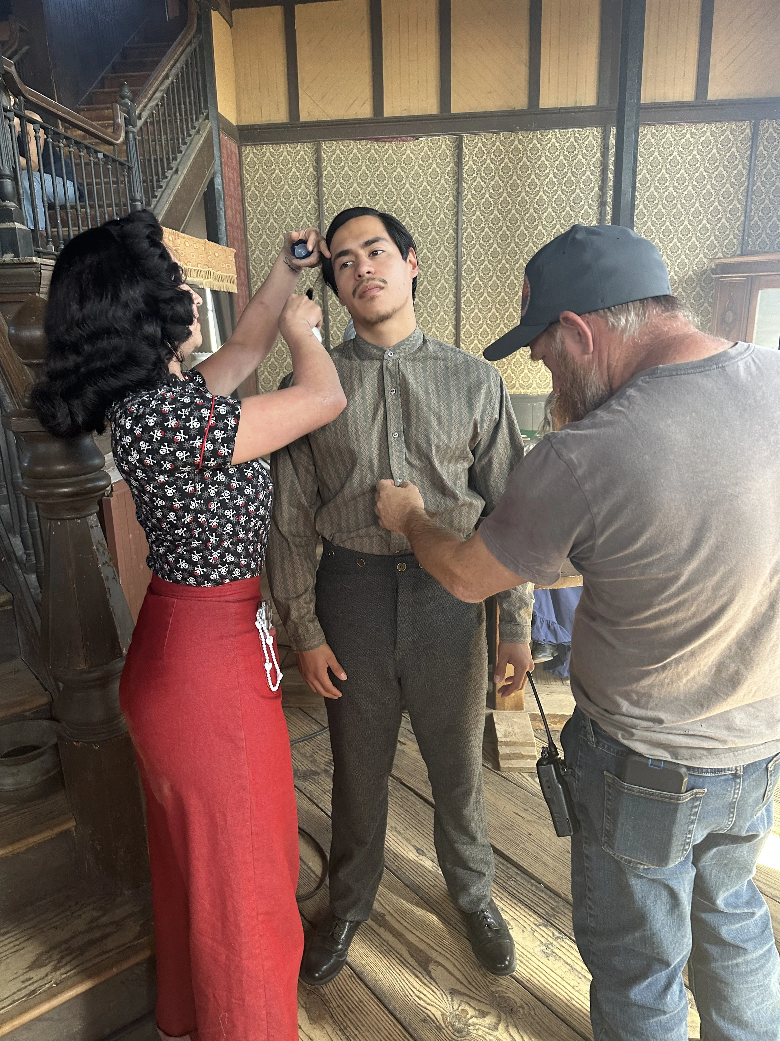 On set of a film or television show, a man is having his costume adjusted while a woman with dark, wavy hair and a red skirt looks on, and another crew member with a beard and cap helps with the adjustments, all inside a vintage-style room with patte