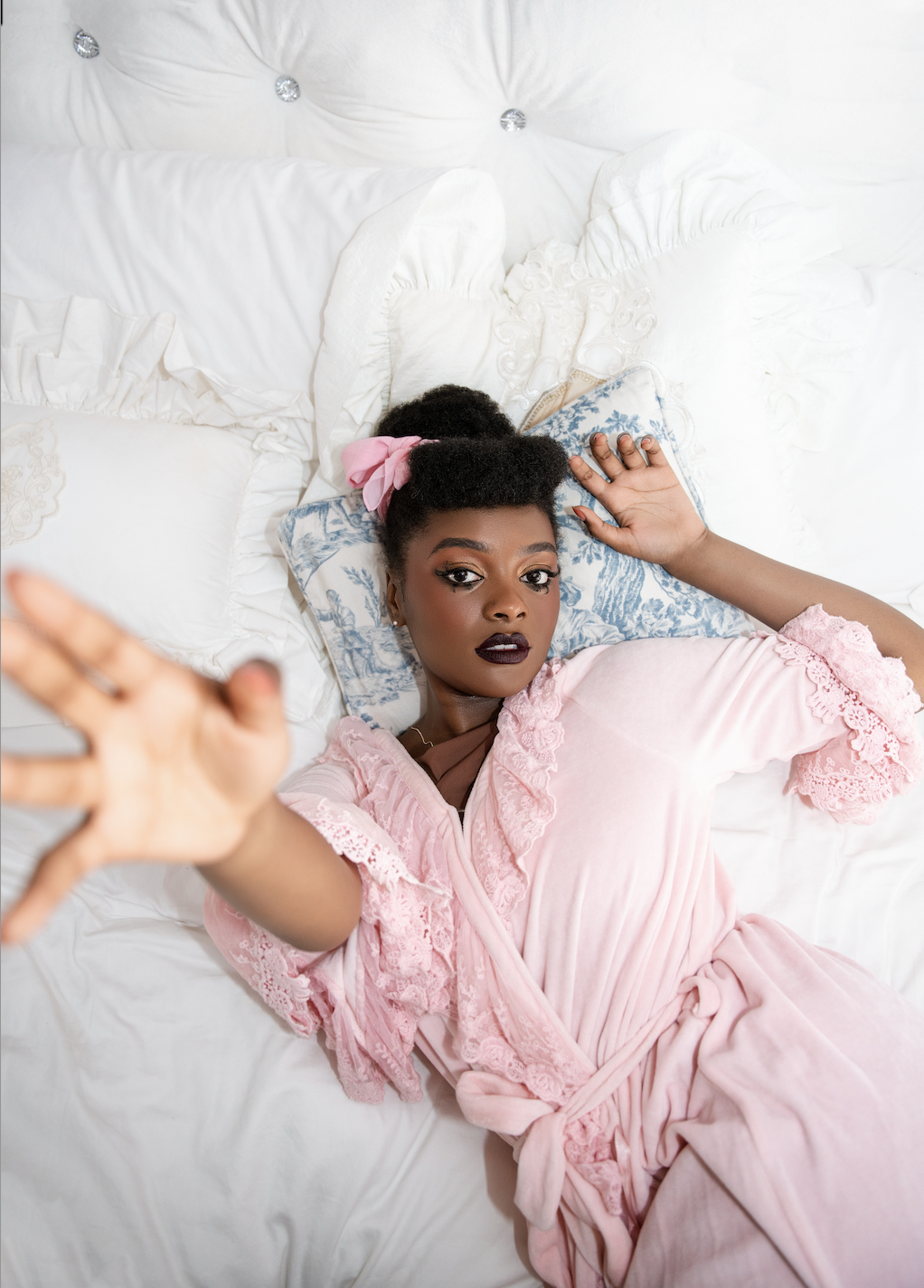 A woman with dark hair styled in an updo with a pink bow, lying on a white bed with pillows. She is wearing a pink nightgown with lace details and reaching out toward the camera with a serious expression.