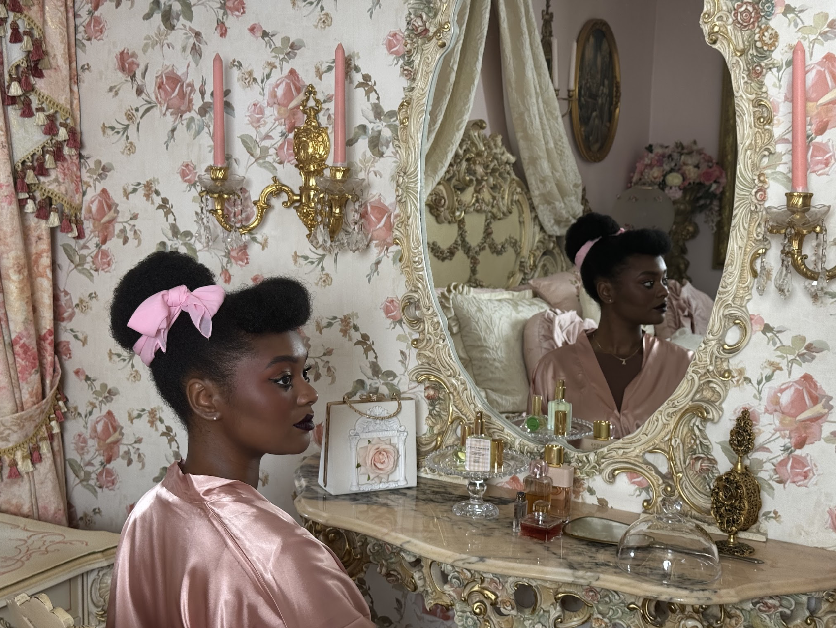 A woman with dark skin, styled in vintage fashion with a pink satin robe and a large pink bow in her hair, sits at a vanity in a luxurious, vintage-style bedroom. She looks at her reflection in an ornate mirror surrounded by perfume bottles and decor