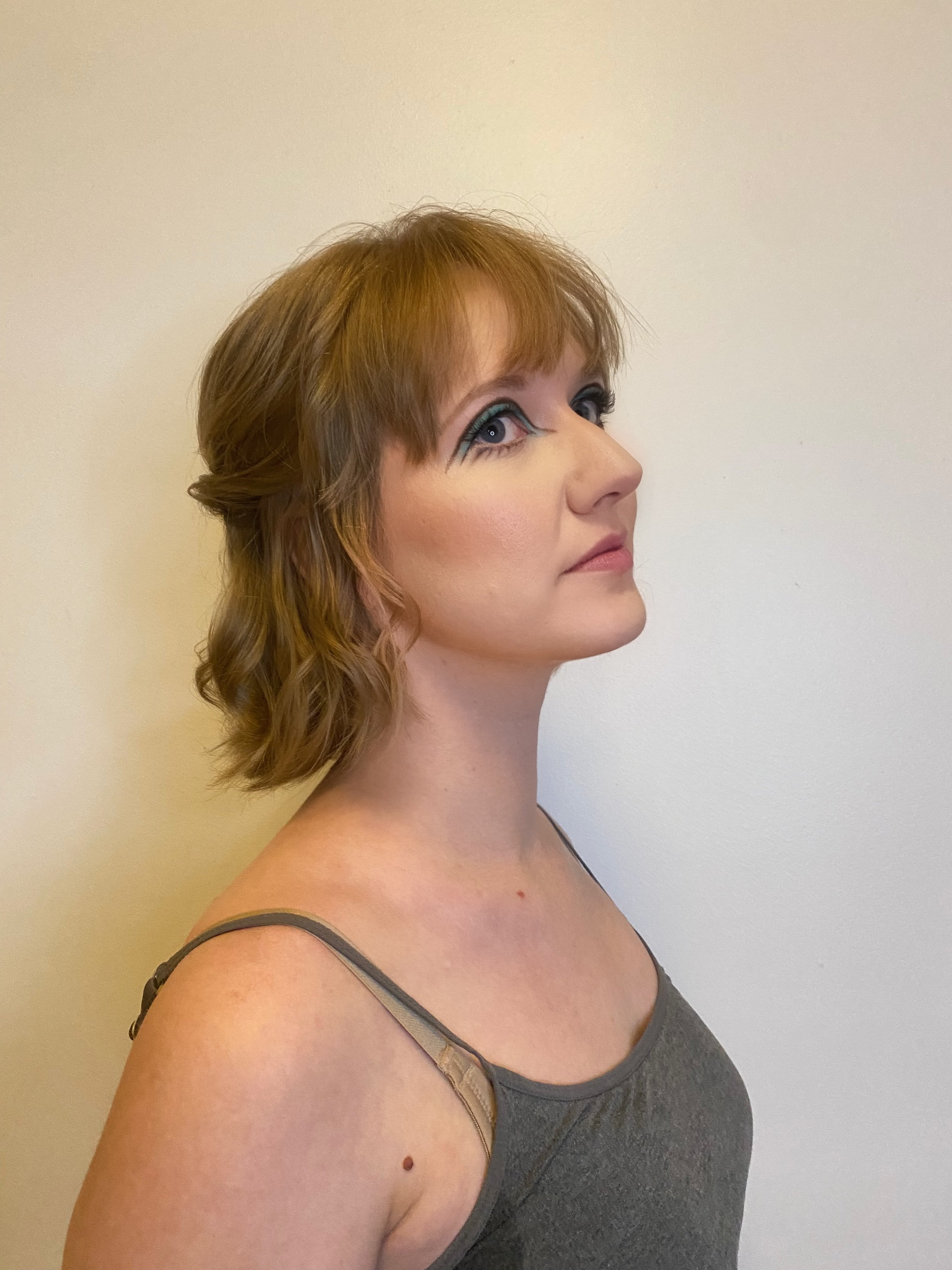 A woman with short, wavy auburn hair, wearing a gray spaghetti strap top, looking upward against a plain off-white wall.