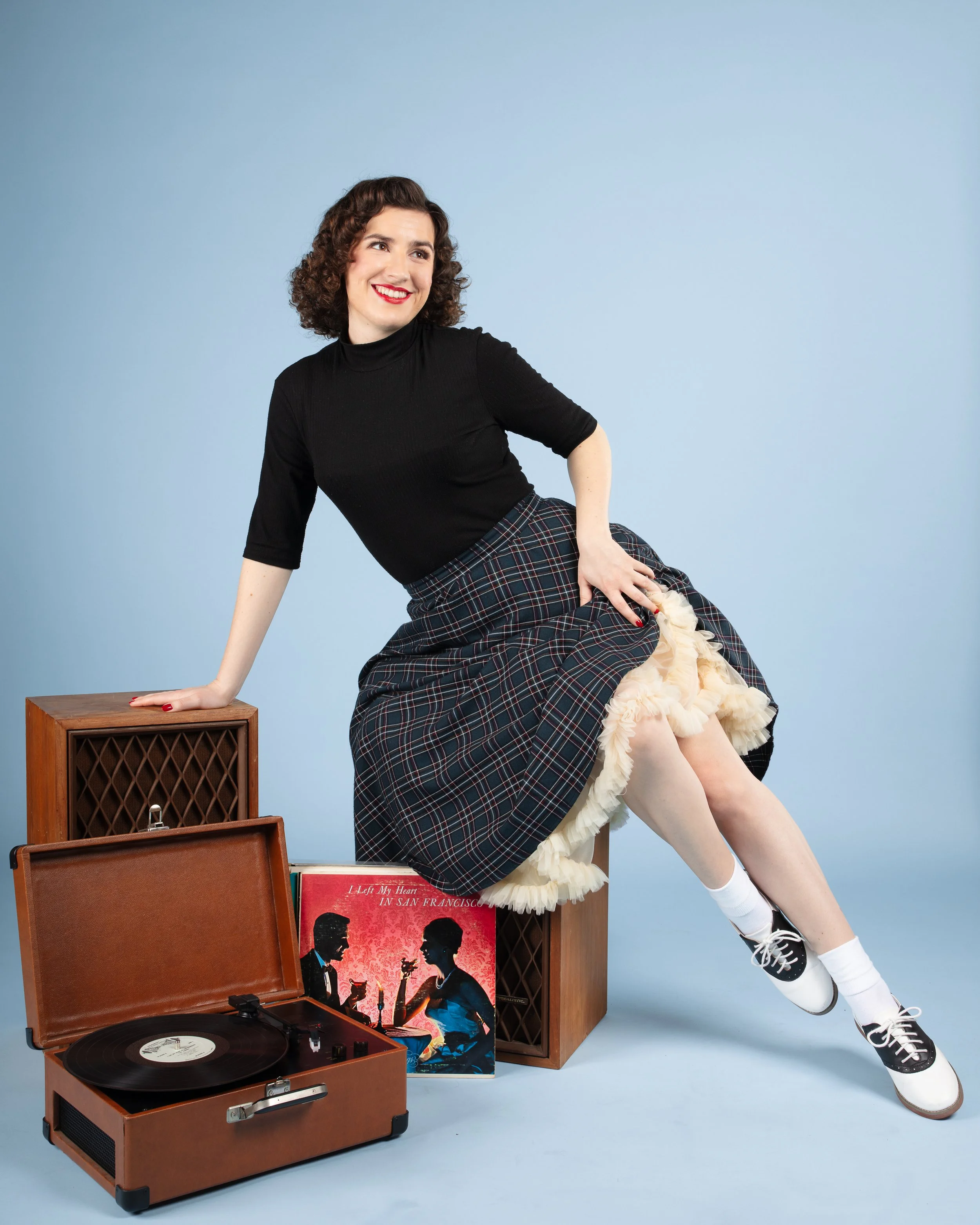 A woman with curly brown hair, wearing a black top and plaid skirt, sitting on a wooden crate against a light blue background. She is smiling and holding up her skirt to reveal a petticoat. There is a vintage record player and a vinyl record with a cover titled "I Left My Heart in San Francisco" in front of her.