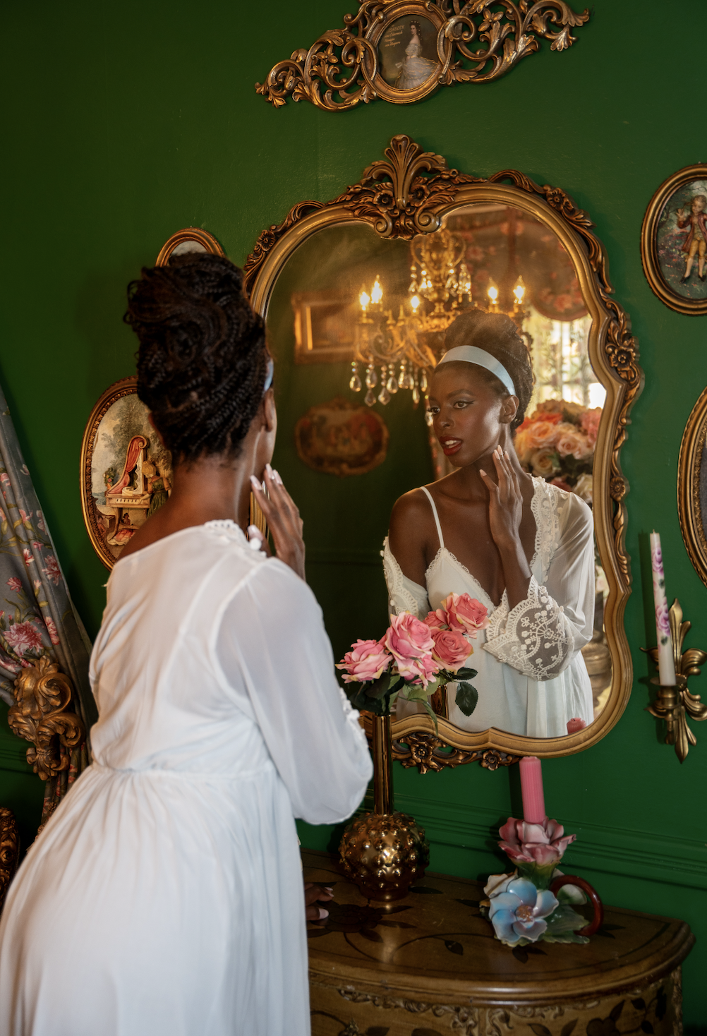 A woman in a white dress looks at herself in an ornate mirror, touching her face, with a bouquet of pink roses on a vintage dresser. The room has green walls and vintage framed portraits, with a chandelier reflecting in the mirror.