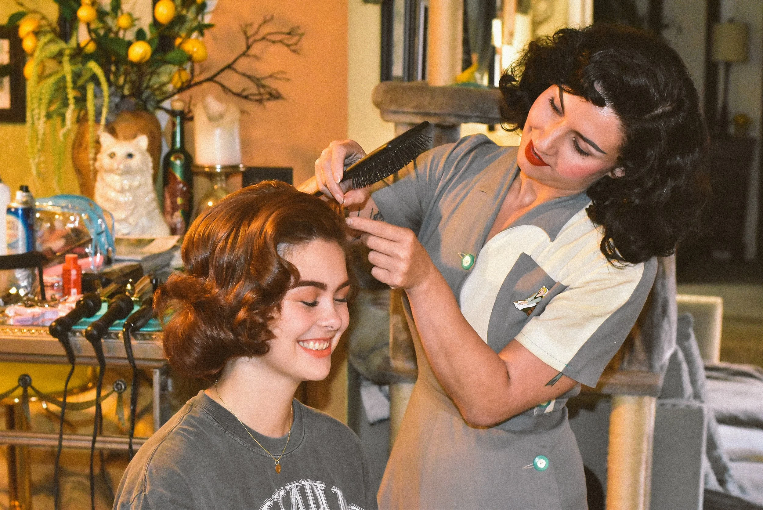 A woman with black curly hair and red lipstick is styling another woman with short, wavy brown hair in a vintage hairdo. The woman being styled is smiling with her eyes closed. They are in a cozy room decorated with a ceramic cat figurine, flowers, a