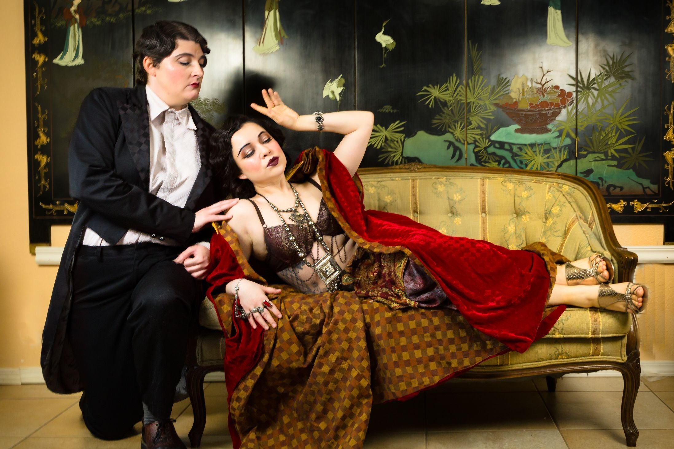 Two women in gothic and vintage-inspired clothing, one sitting and the other lying on a vintage yellow sofa with a black and gold Asian-style screen behind them.