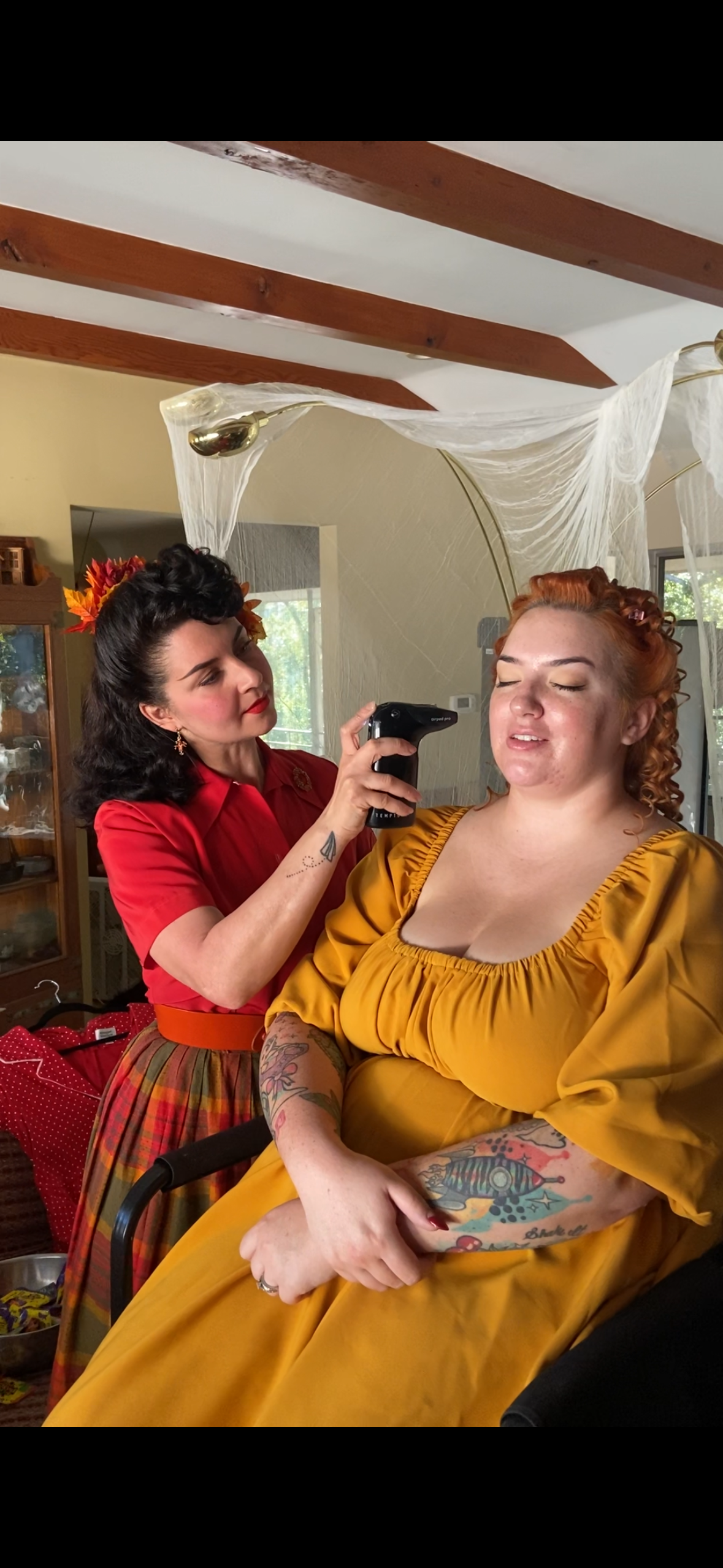 A woman in a yellow dress with tattoos on her arms is having her temperature taken with a forehead thermometer by another woman dressed in vintage style with a red shirt, floral headband, and a plaid skirt, in a cozy room decorated with spider web curtains.