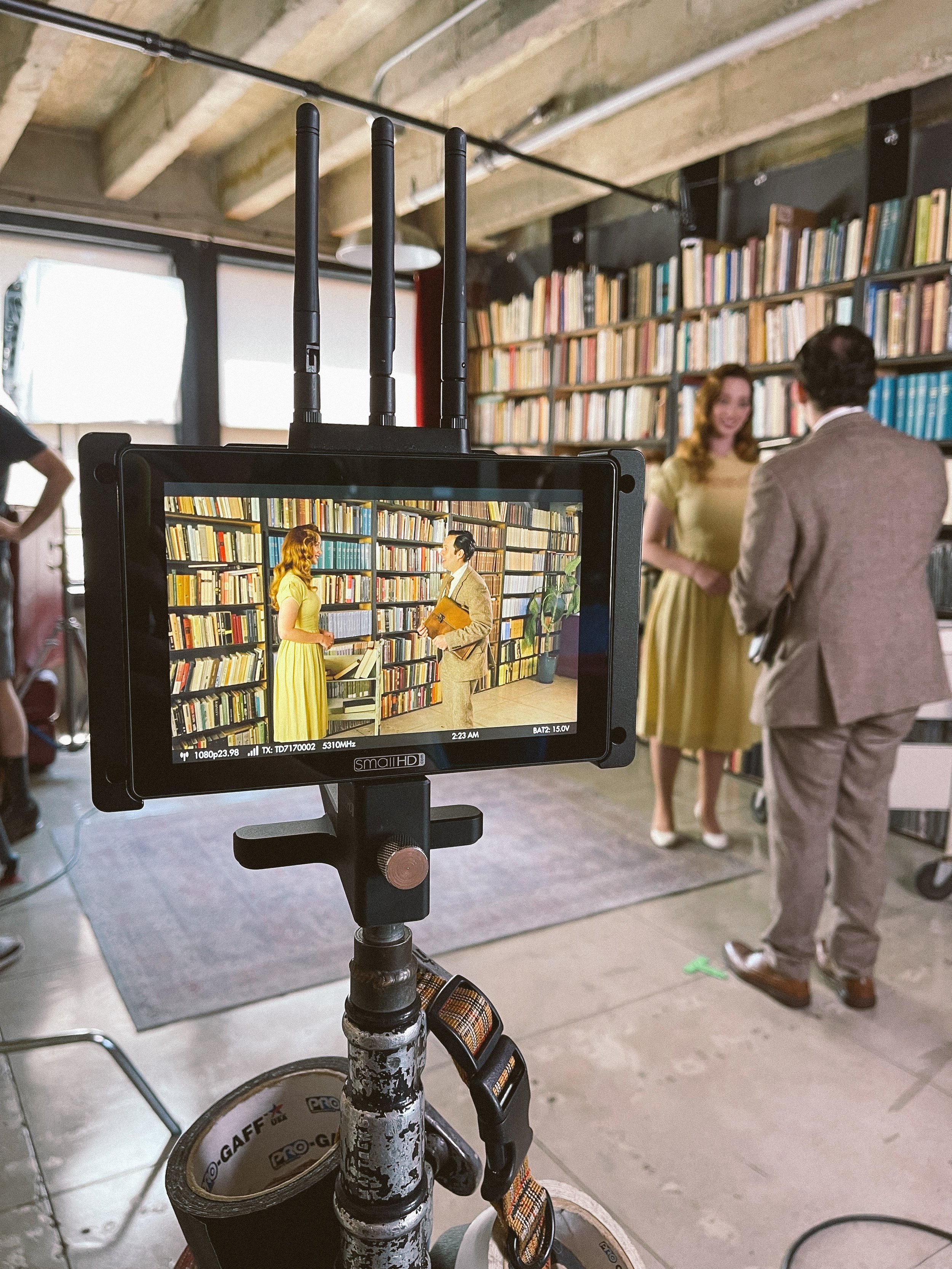 People on a film set in a library, with a red-haired woman and a man with dark hair in a beige suit having a conversation, seen through a monitor capturing the scene. The scene is staged in front of a large bookshelf filled with books.