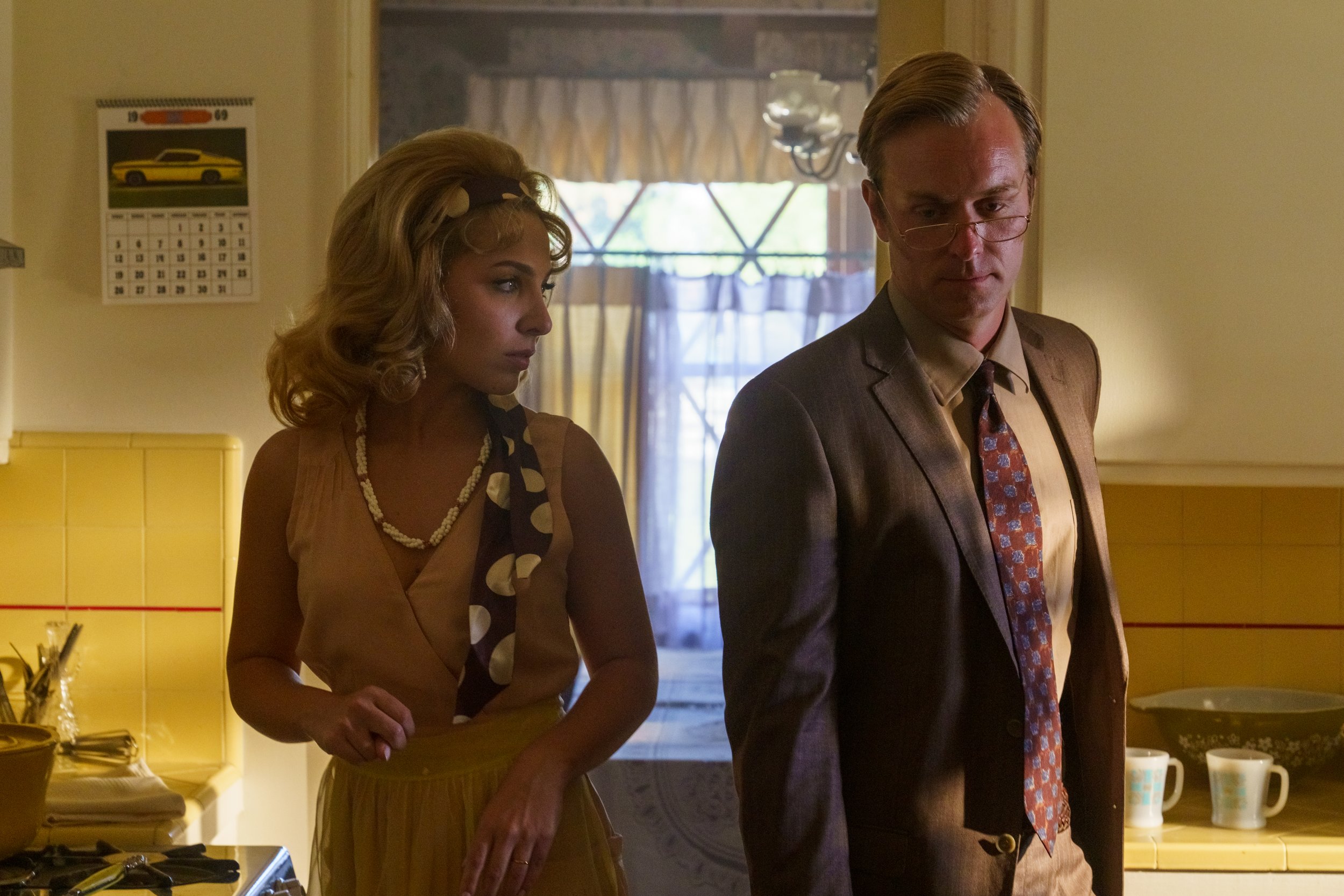 A woman with curly blonde hair and a man wearing glasses and a suit are standing in a kitchen, looking down with serious expressions.