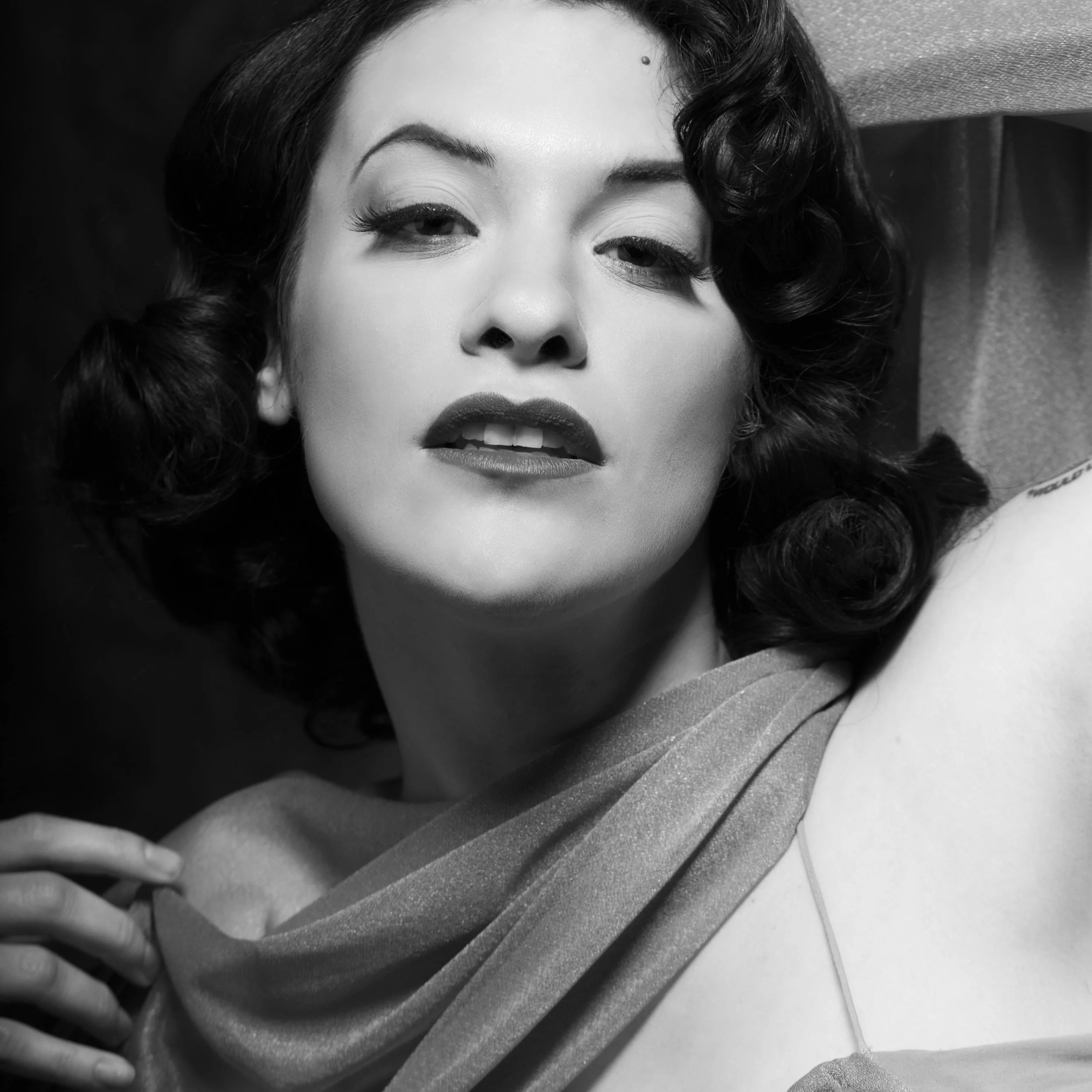 Black and white portrait of a woman with vintage hairstyle, bold lips, and makeup, posing with a confident expression, wearing a draped fabric around her shoulder.