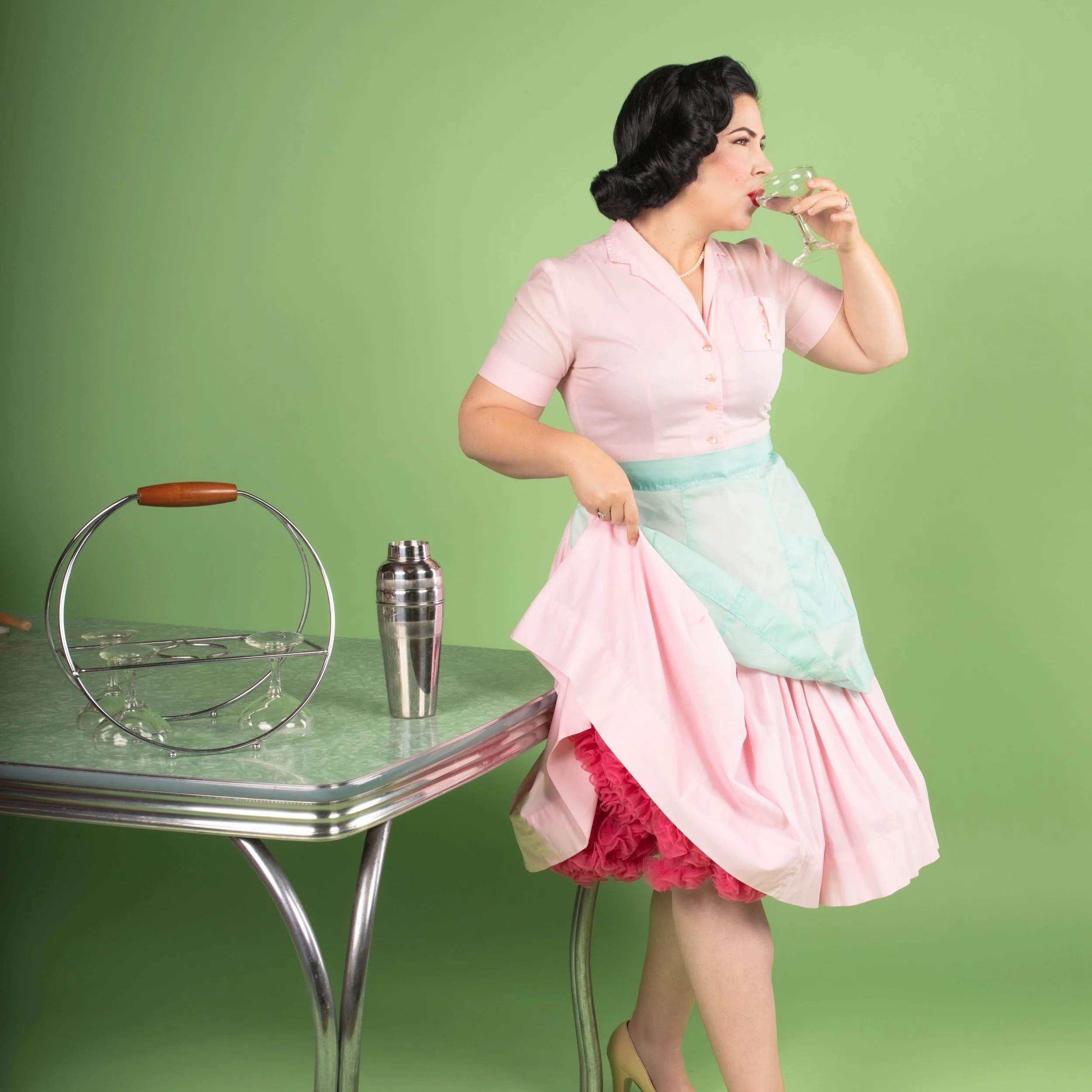 A woman with dark, styled hair and wearing a retro pink dress with pastel apron, standing by a vintage green table, drinking from a cocktail glass, with a stainless steel shaker and cocktail strainer on the table, against a green background.