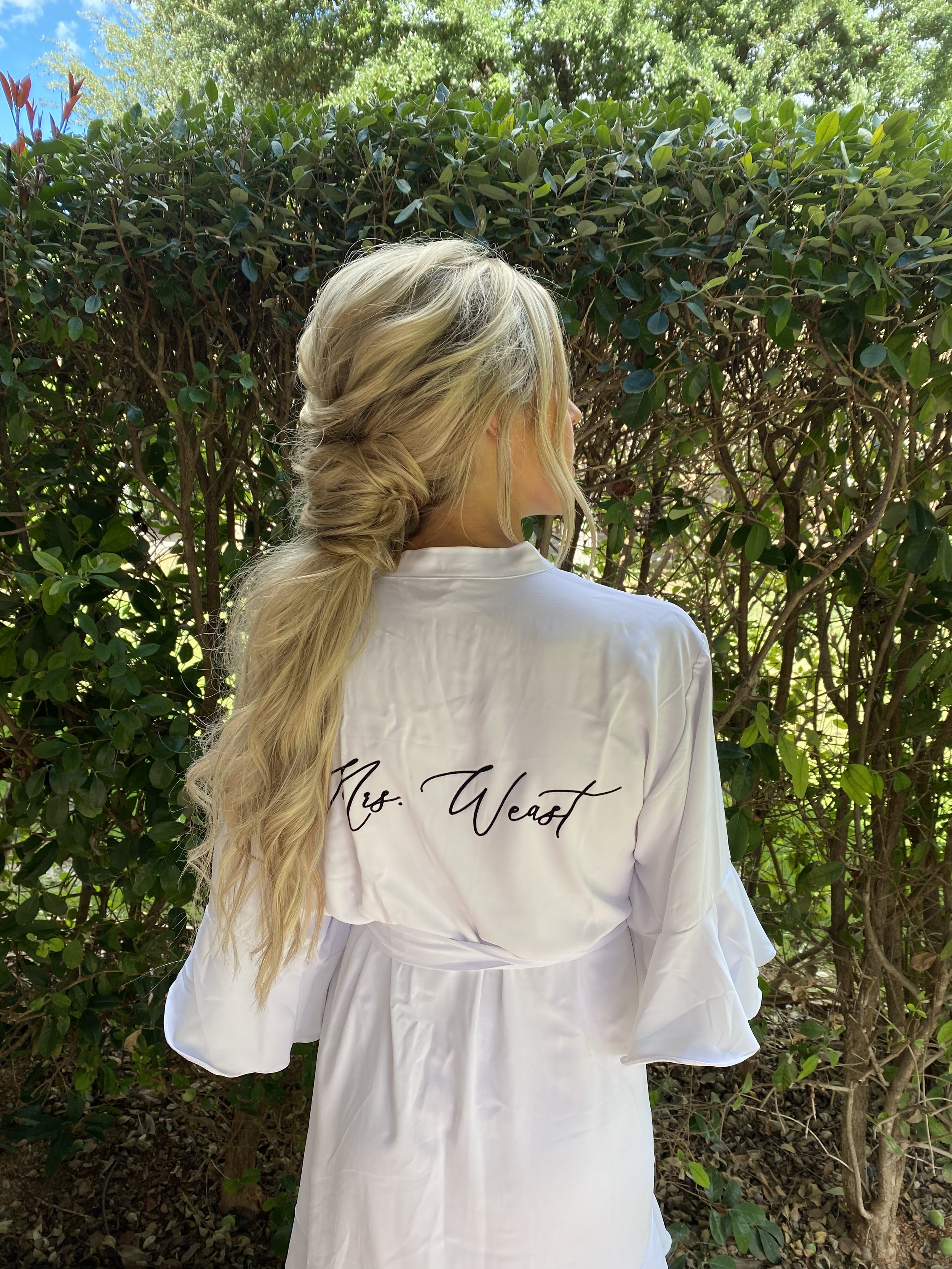 Woman with long blonde hair styled in loose waves and a braided side, wearing a white satin robe with 'Mrs. West' embroidered on the back, standing outdoors in front of green bushes.