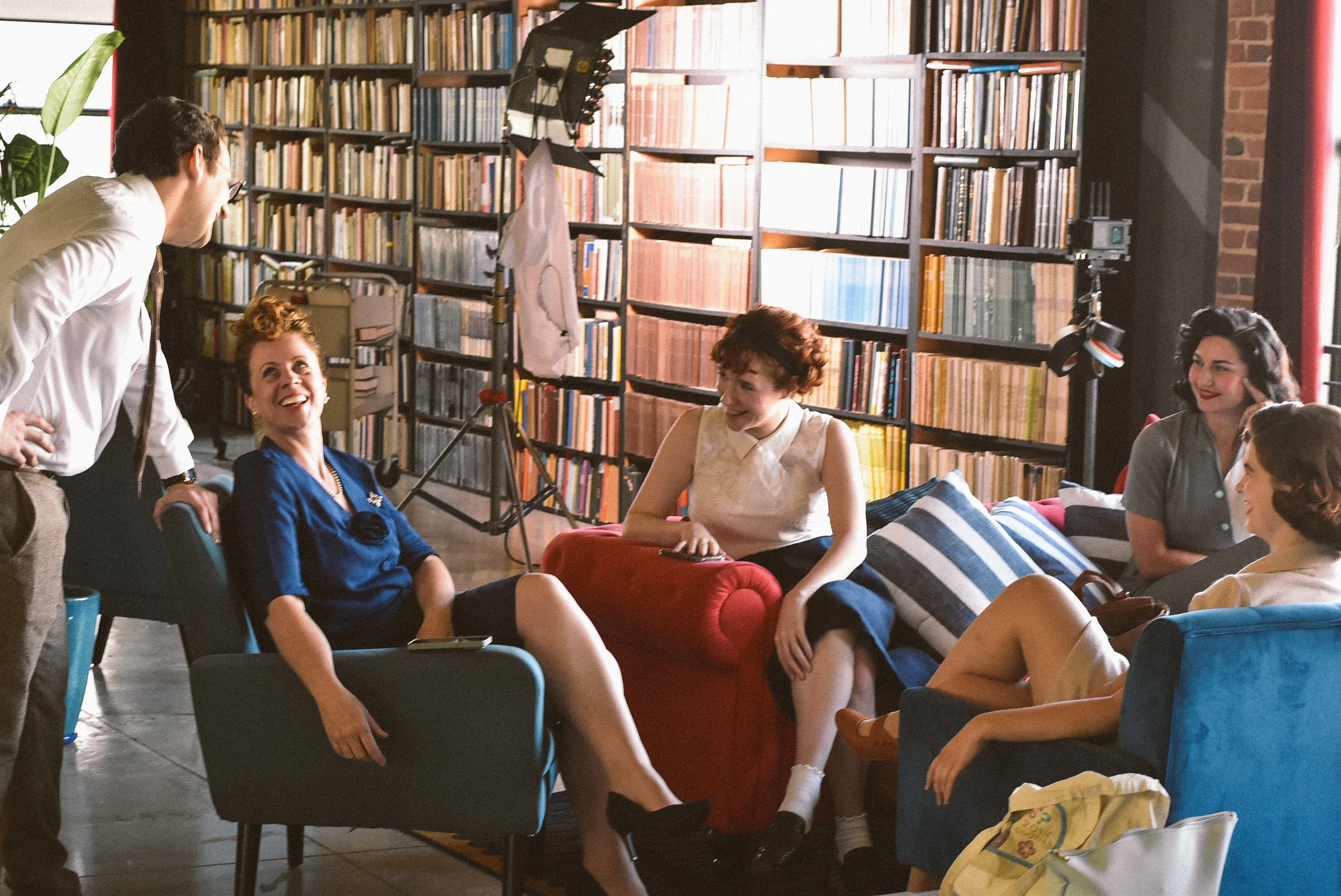 A group of six people having a lively discussion or interview in a cozy room with bookshelves. They are seated on various colored chairs and couches, smiling and engaging with each other. There is professional filming equipment, including a camera an