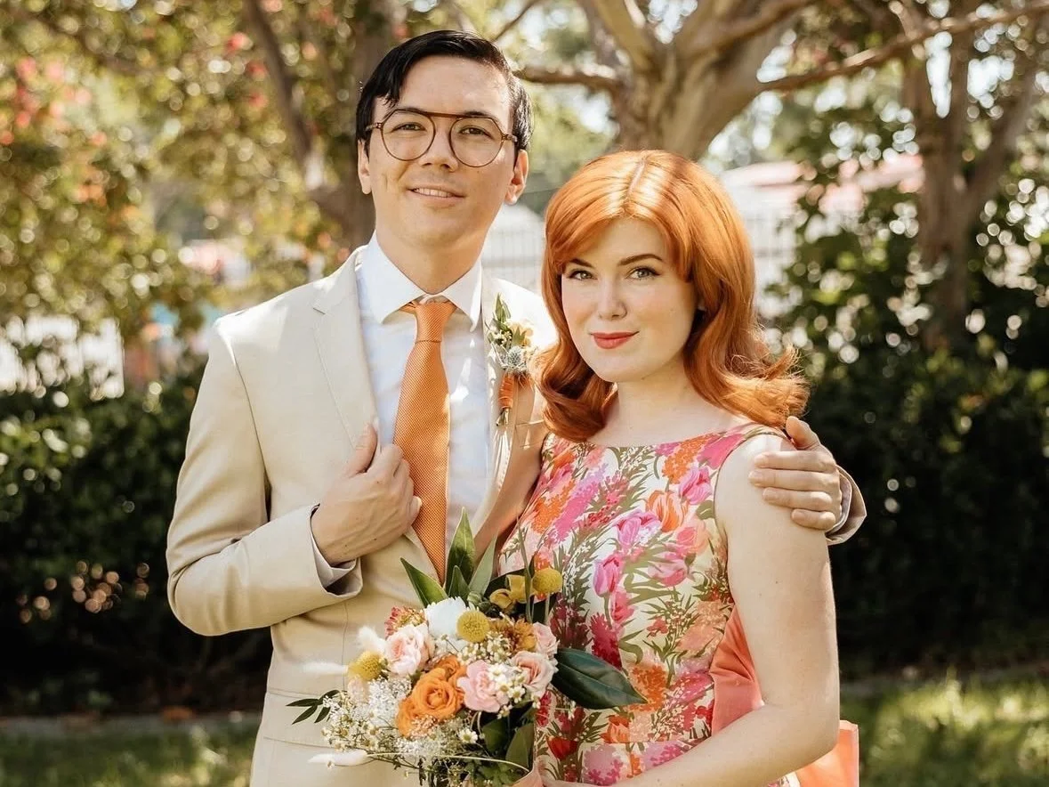 A young couple posing outdoors, dressed in wedding attire with the man in a cream suit and orange tie, and the woman in a floral dress holding a bouquet of flowers. They are standing in front of a tree with green foliage.