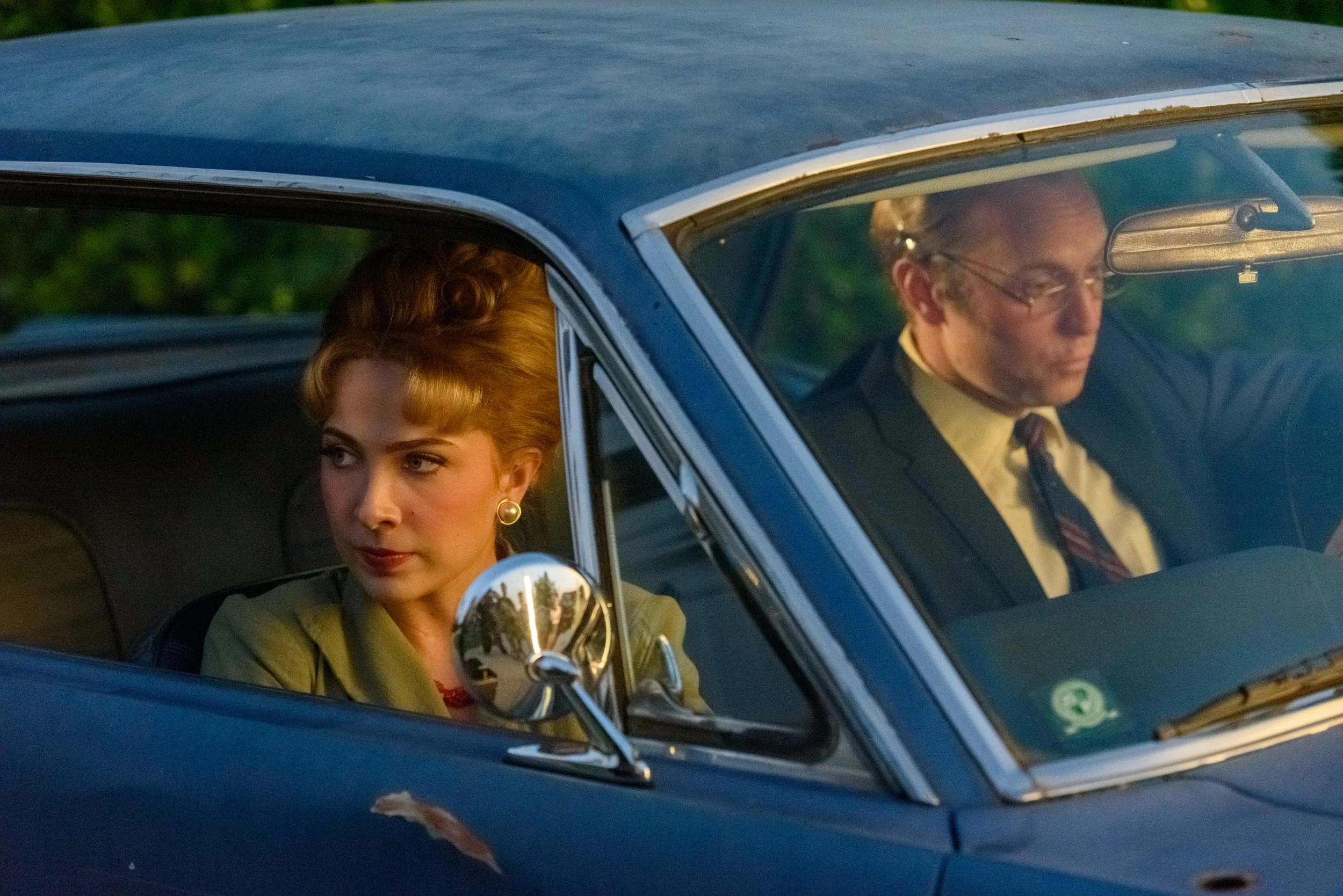 A woman and a man sit inside a vintage blue car, with the woman in the driver's seat and the man in the passenger seat, both looking serious.