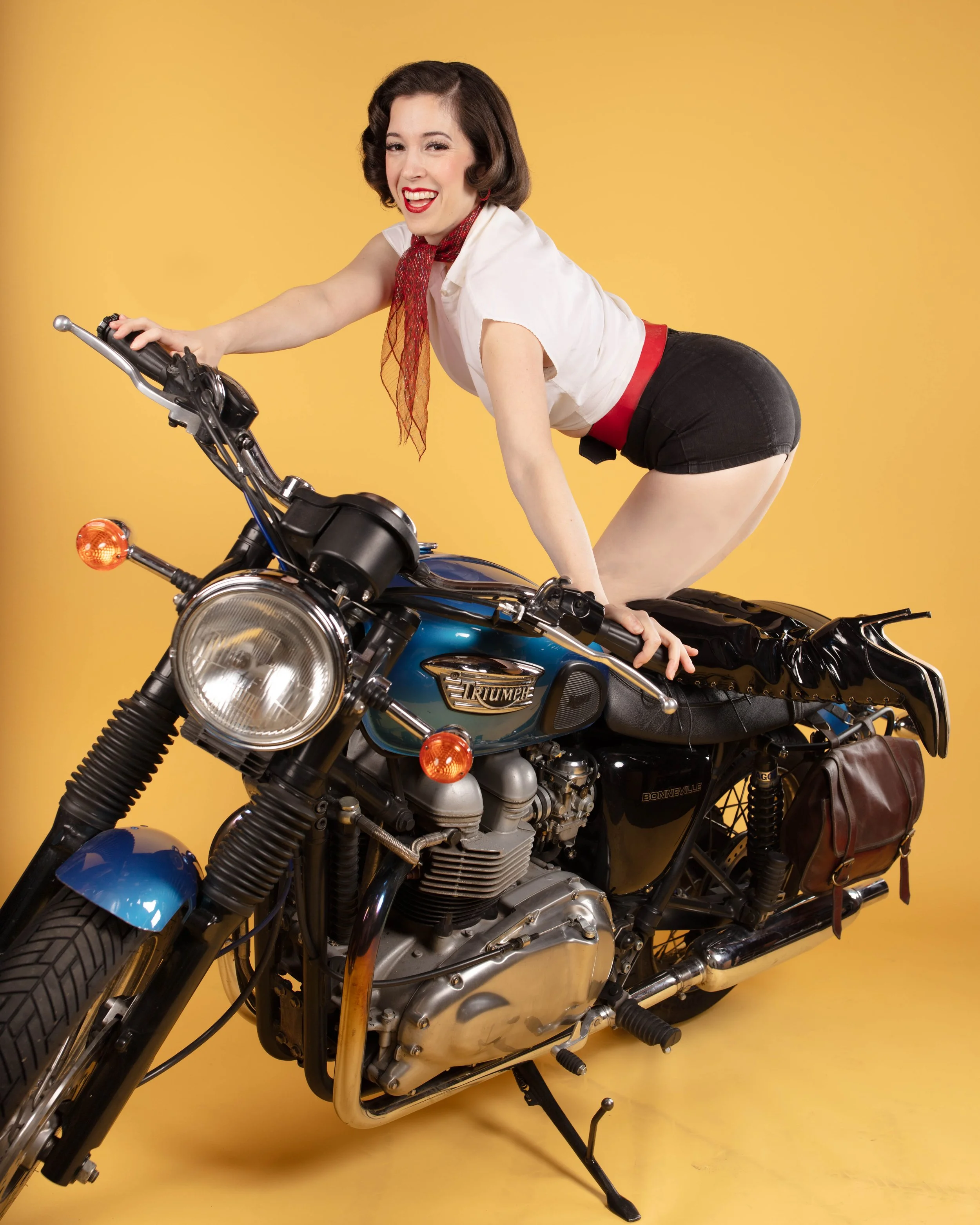 A woman with dark hair, wearing a white top, black shorts, and red accessories, is sitting on a blue Triumph Bonneville motorcycle against a yellow background.