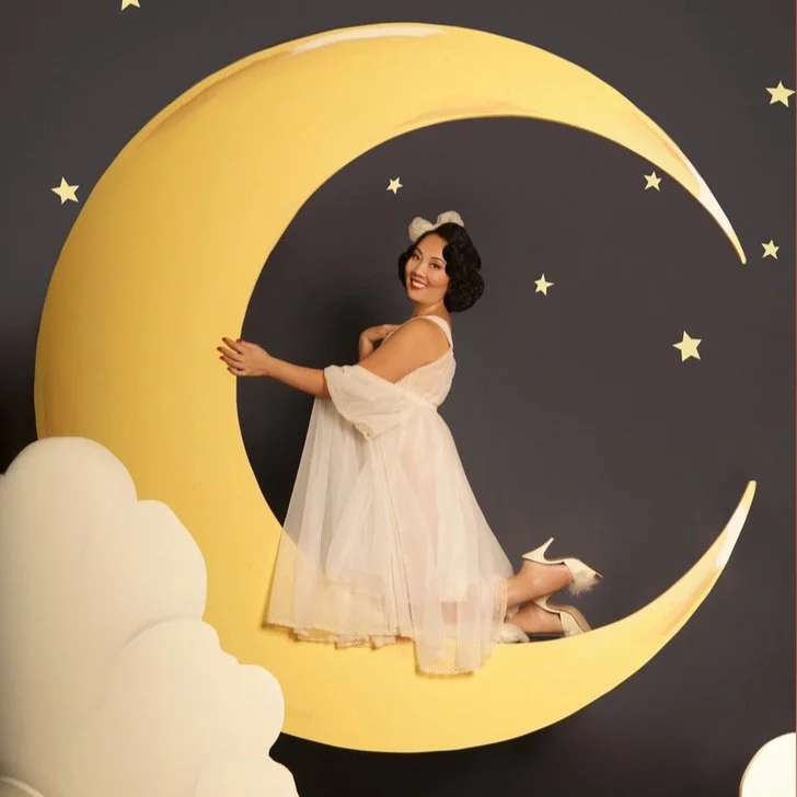 A woman dressed in a white dress and wearing a white headband with a bow, smiling while kneeling inside a large yellow moon cutout with a dark background decorated with small stars and clouds.
