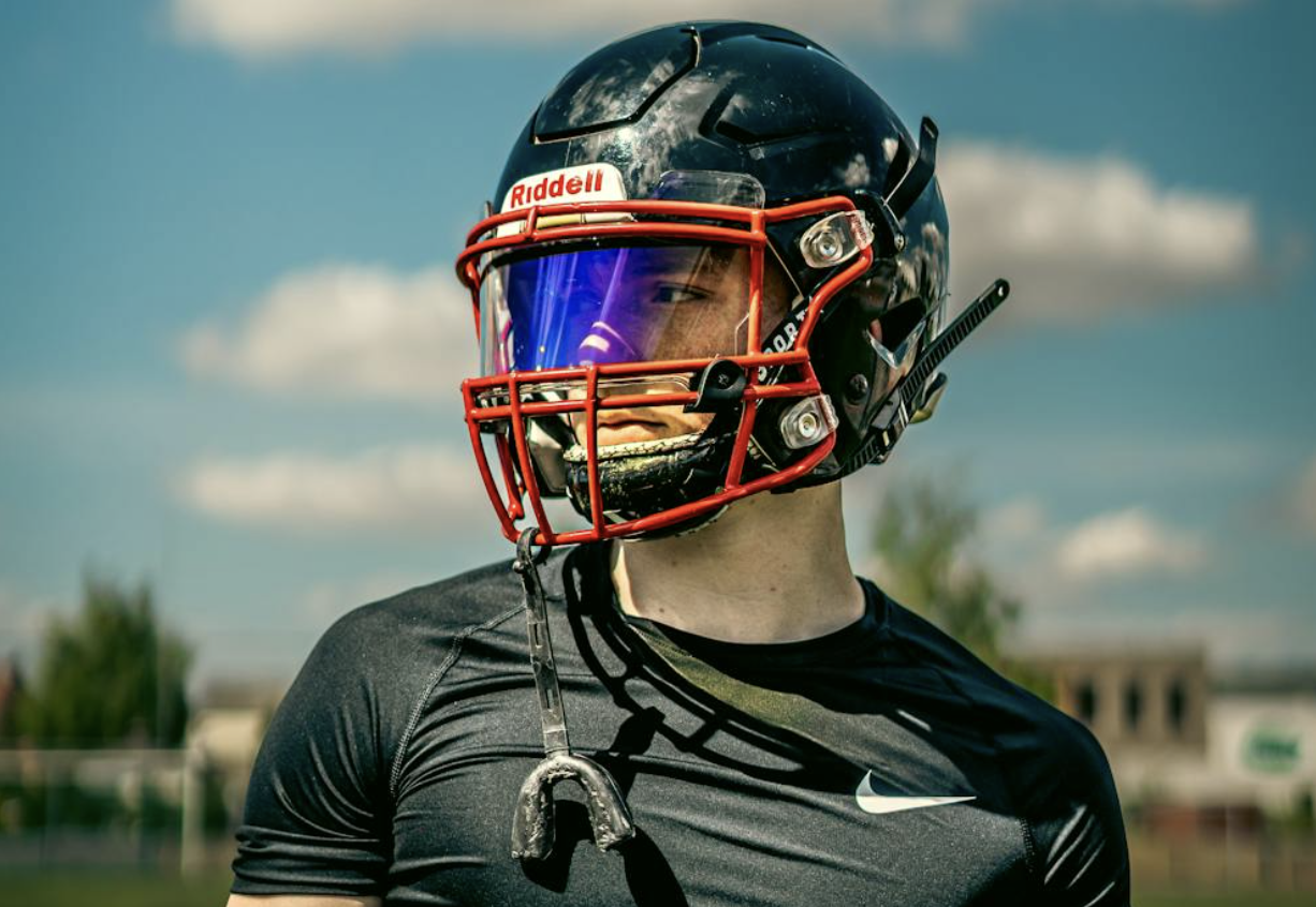 A youth football player looks toward the distance. A mouth guard to protect his teeth dangles from a strap holding it to his helmet.