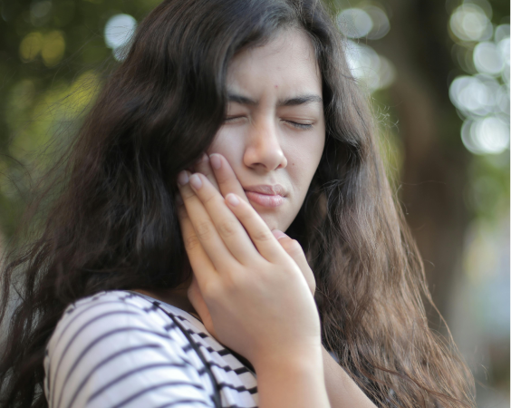 A young woman with long brown wavy hair is holding her jaw with both hands, eyes squeezed shut in obvious pain.