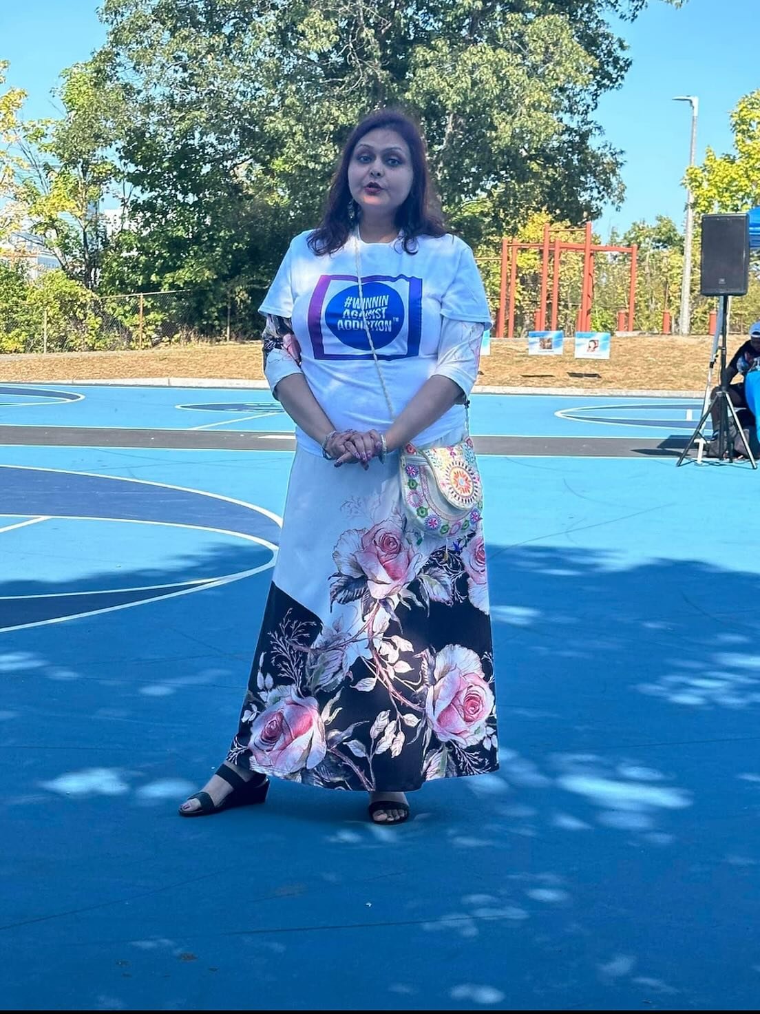 A woman standing on a blue basketball court outdoors during daytime, wearing a white t-shirt with a blue logo and hashtag, and a long floral skirt with pink roses. She has dark hair and appears to be speaking or singing, with trees and a playground i