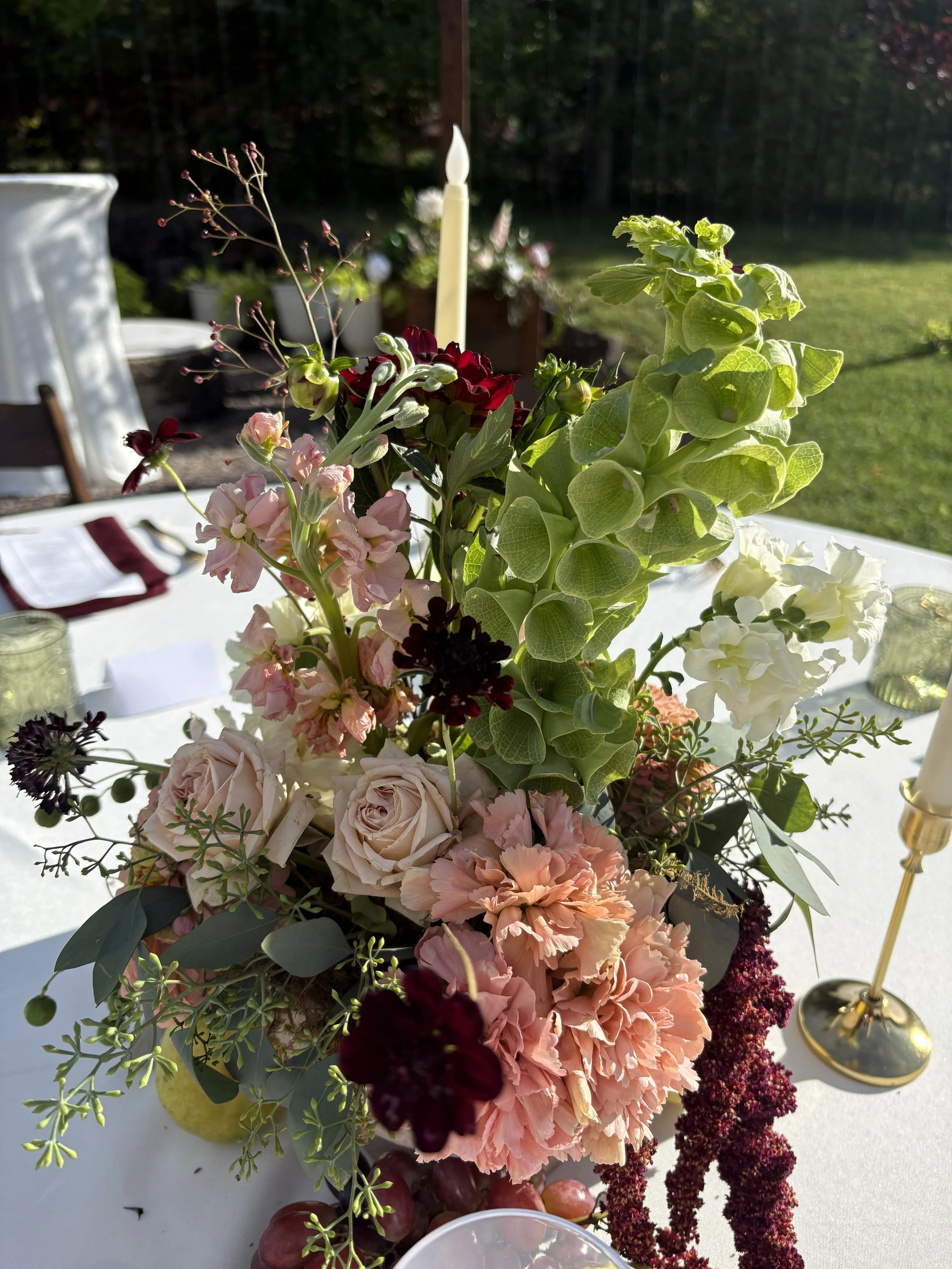 Close-up of a flower arrangement featuring pink roses, red and pink carnations, green foliage, and burgundy amaranthus on a white table, with a lit candle and blurred outdoor background.