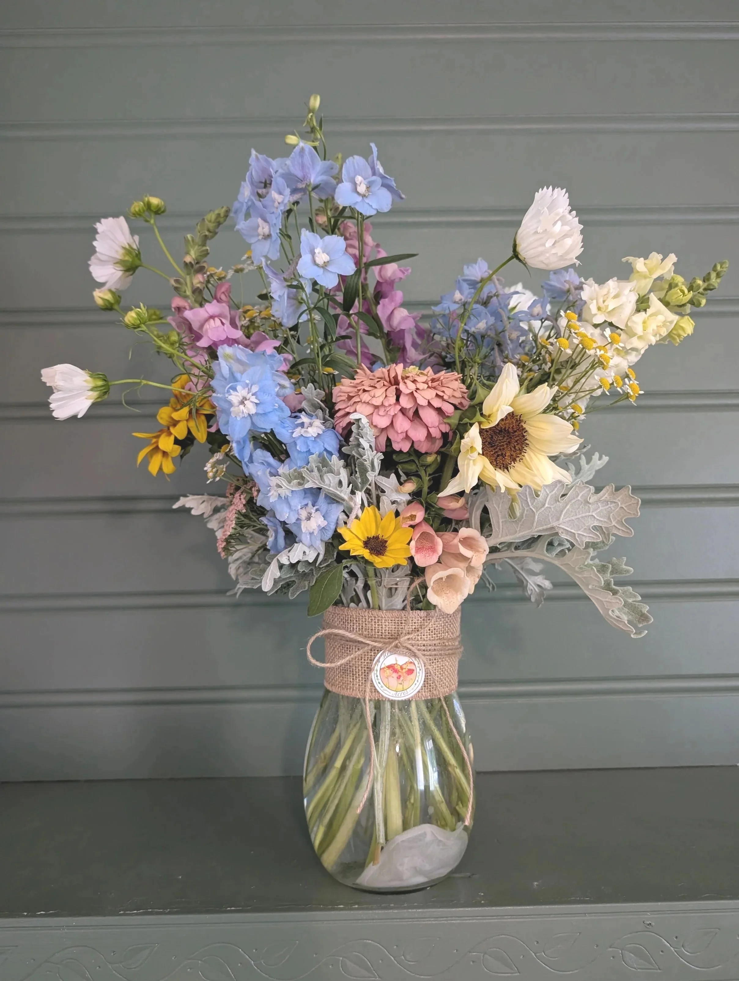 A bouquet of colorful flowers in a glass vase, wrapped with burlap and ribbon, placed on a black surface against a gray paneled wall.