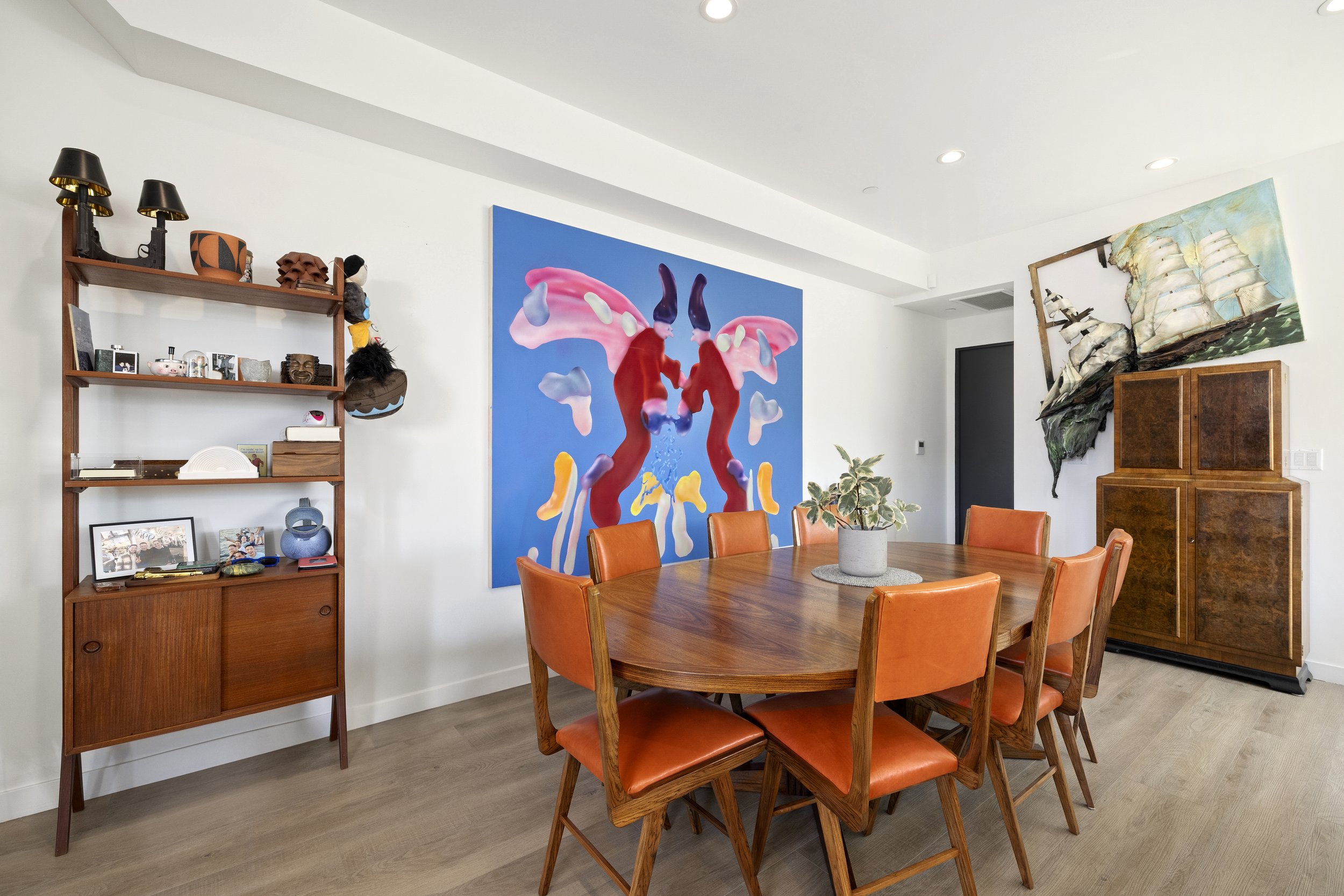 A dining room with a wooden table surrounded by orange chairs, a potted plant in the center, two large colorful abstract paintings on white walls, and a wooden bookshelf on the left with various decorative items.