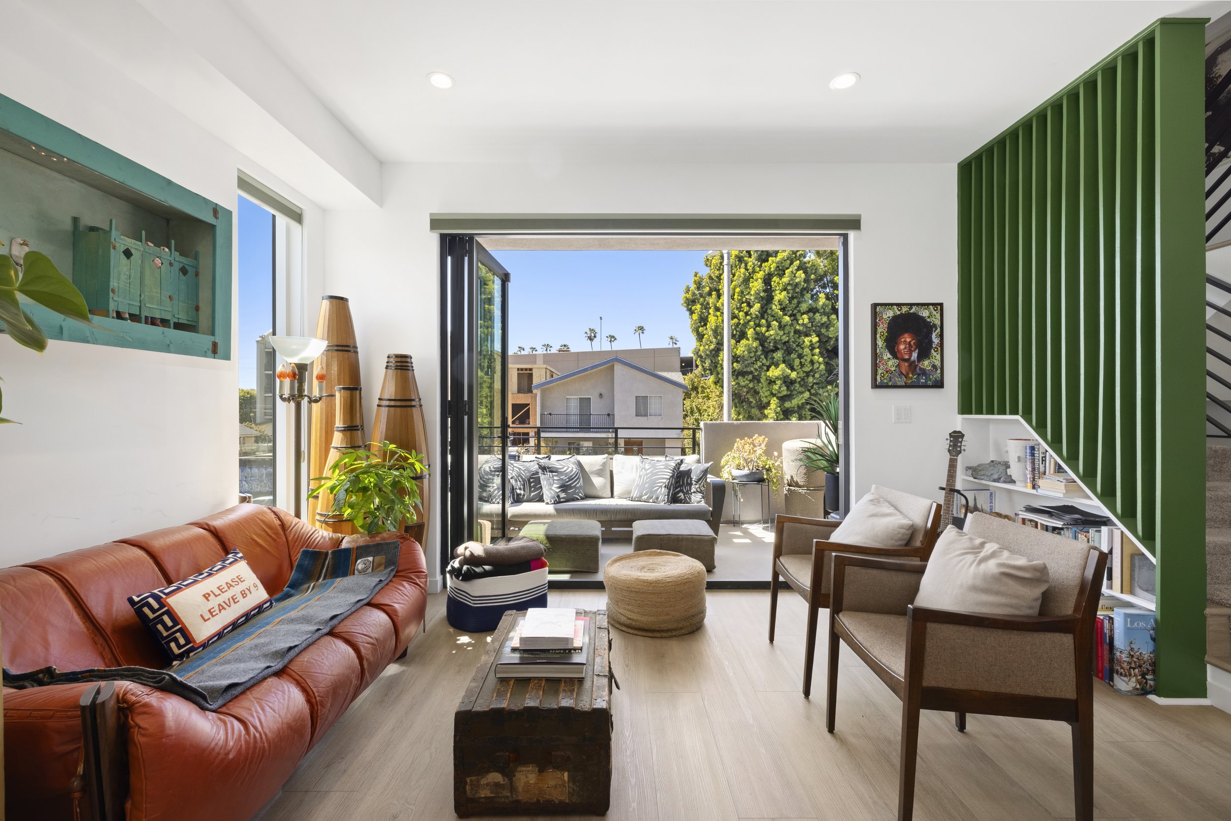 Living room with leather sofa, wooden chairs, green bookshelf, and open balcony door showing outdoor seating and trees