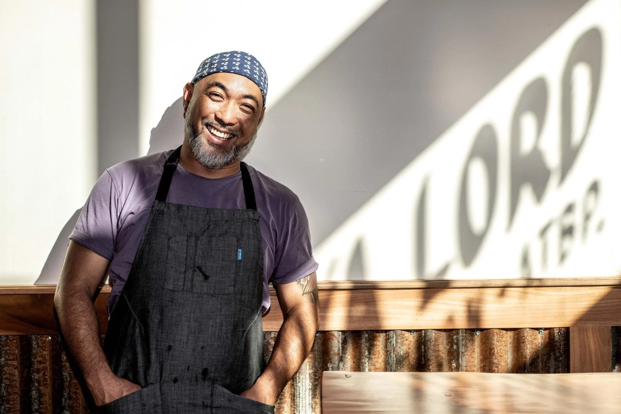 Smiling man wearing a blue bandana, purple t-shirt, and black apron standing indoors near a wall with a logo shadow.