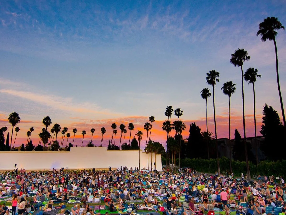 A large outdoor movie screening event at sunset with many people seated on blankets and lawn chairs, surrounded by tall palm trees.