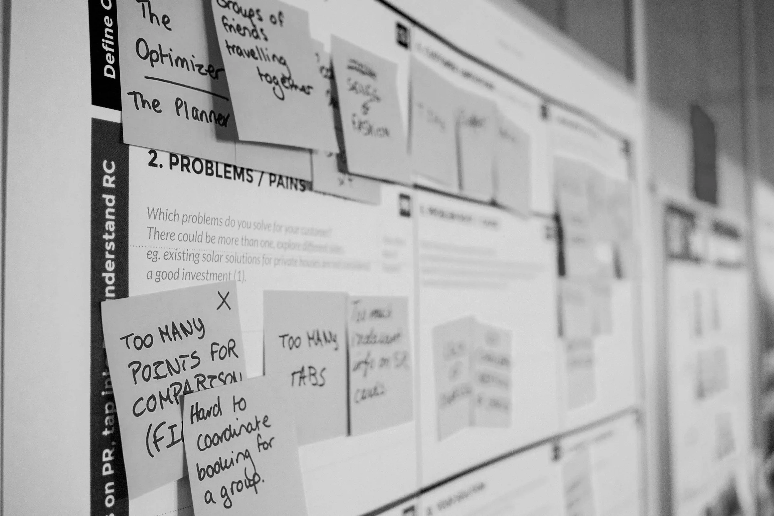 Black and white photo of a bulletin board with notes and papers from a website UX problem-solving workshop with handwritten notes mentioning points like 'Too many points for comparison' and 'Hard to coordinate for a group.'