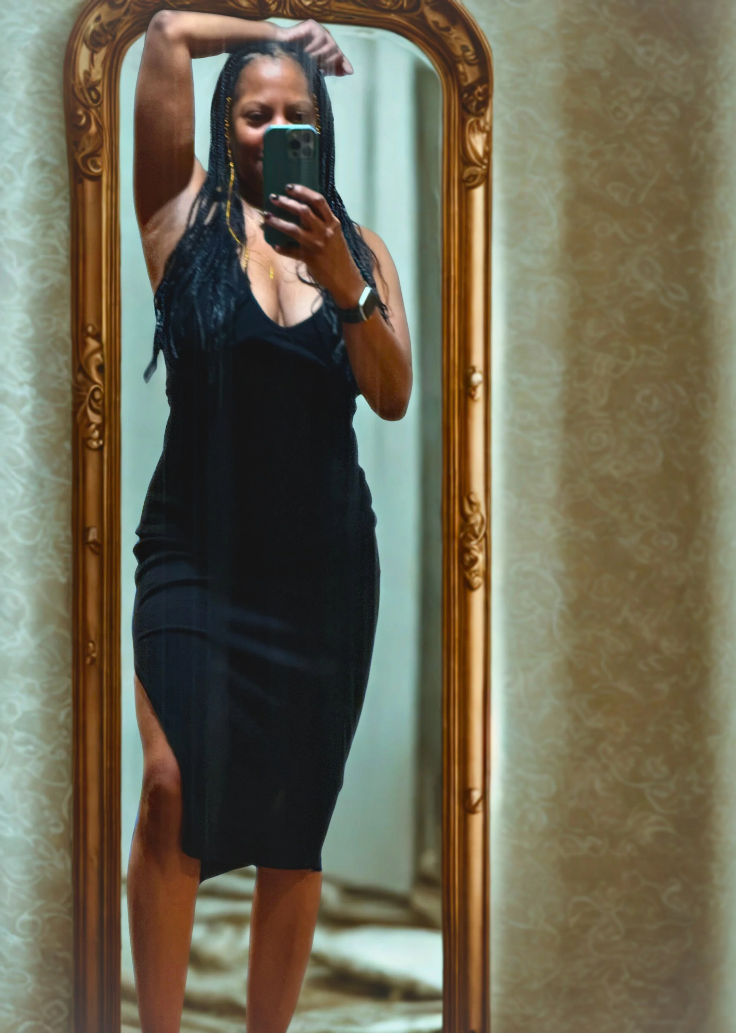 A woman taking a mirror selfie in a black dress with a slit, posing with one arm raised above her head. She has long braided hair and is standing in front of a wooden-framed mirror with patterned wallpaper behind her.