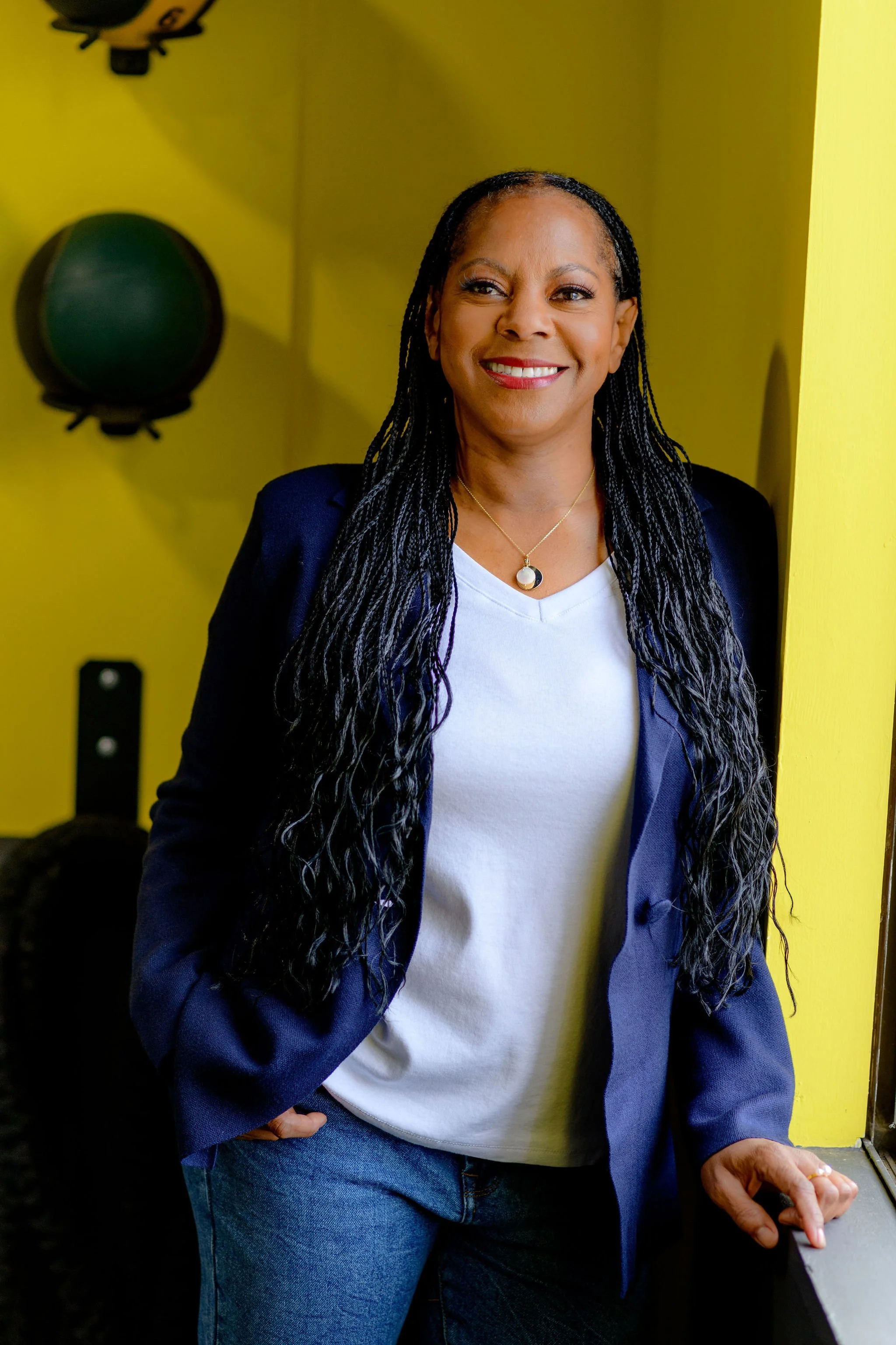 A smiling woman in a navy blazer and white shirt standing near a window with yellow walls and gym equipment in the background.