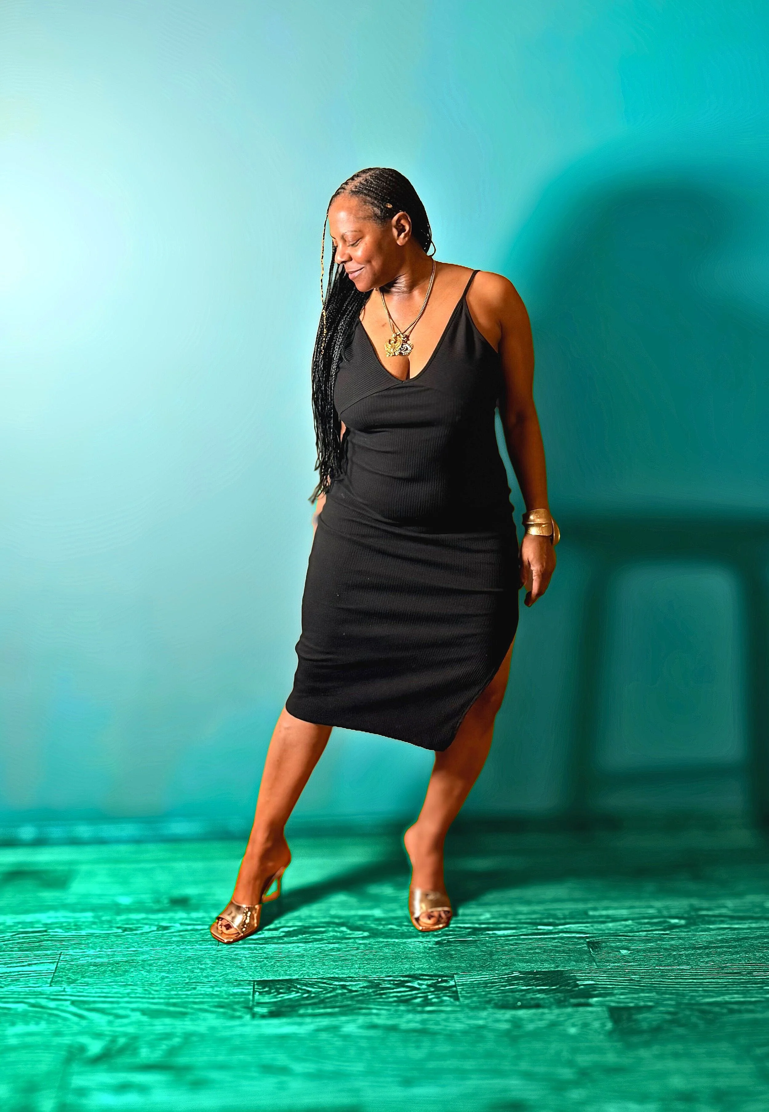 A woman wearing a black sleeveless dress and gold high-heeled shoes, standing against a teal wall and wooden floor, smiling with her head tilted down.