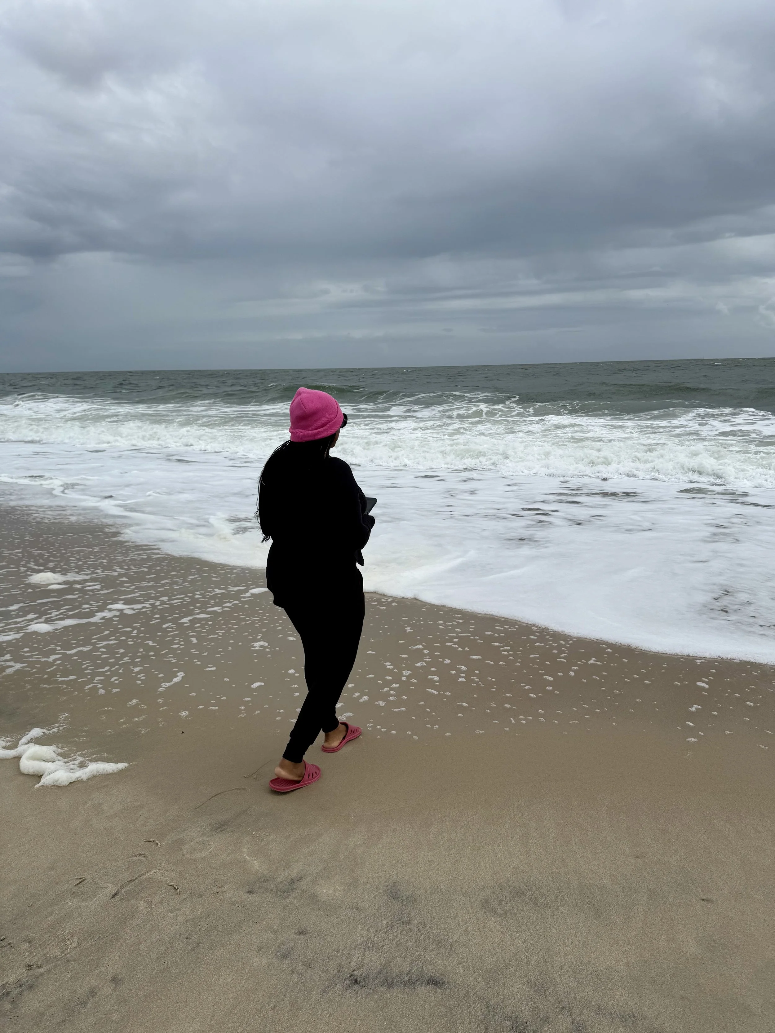 Person in black clothing, pink hat, and pink slippers standing on sandy beach, looking at ocean with waves and cloudy sky.