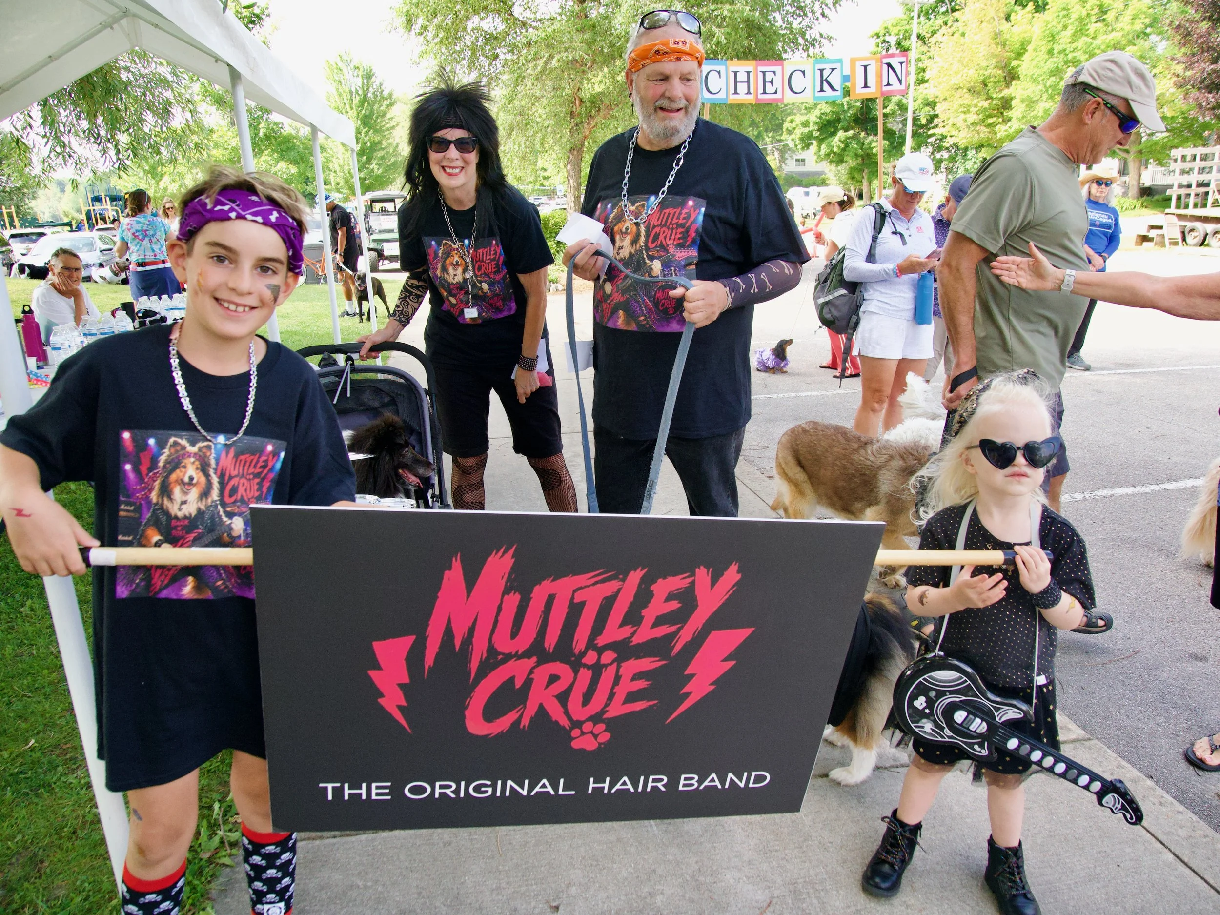 People at an outdoor event with a check-in sign; a young girl smiling with face paint and wearing a bandana, and an older man with a beard wearing a bandana and a cartoon-themed shirt; several people and dogs in the background.