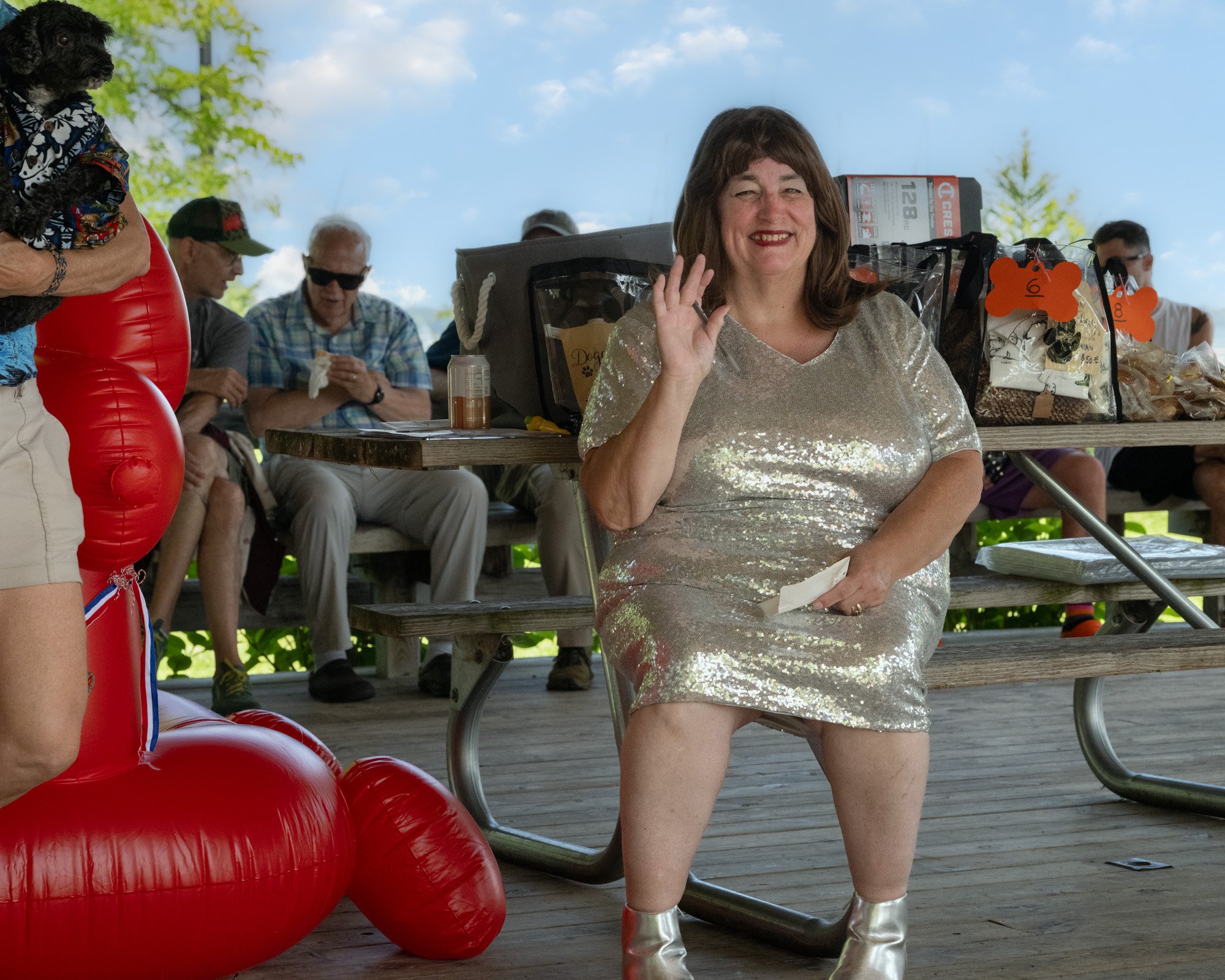 A woman with shoulder-length brown hair wearing a shiny silver dress and silver boots sitting at a picnic table, waving and smiling at the camera, with a woman in a colorful shirt and tan shorts holding a red inflatable object nearby and several peop