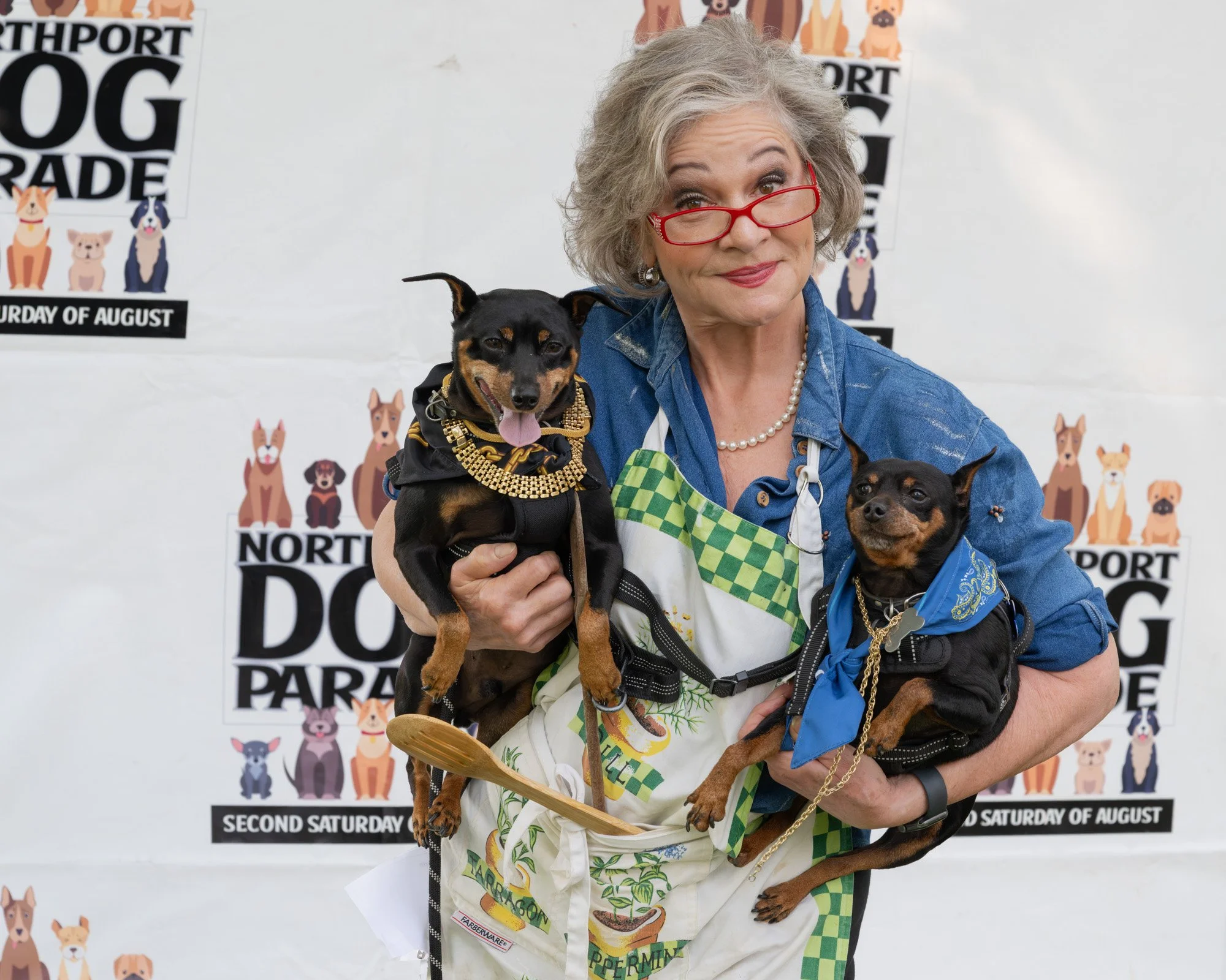 An older woman with gray hair, red glasses, and a pearl necklace holding two small dogs with black and tan fur. The woman is wearing a colorful apron and a denim shirt. The background features a poster with cartoon dogs and text promoting an event in
