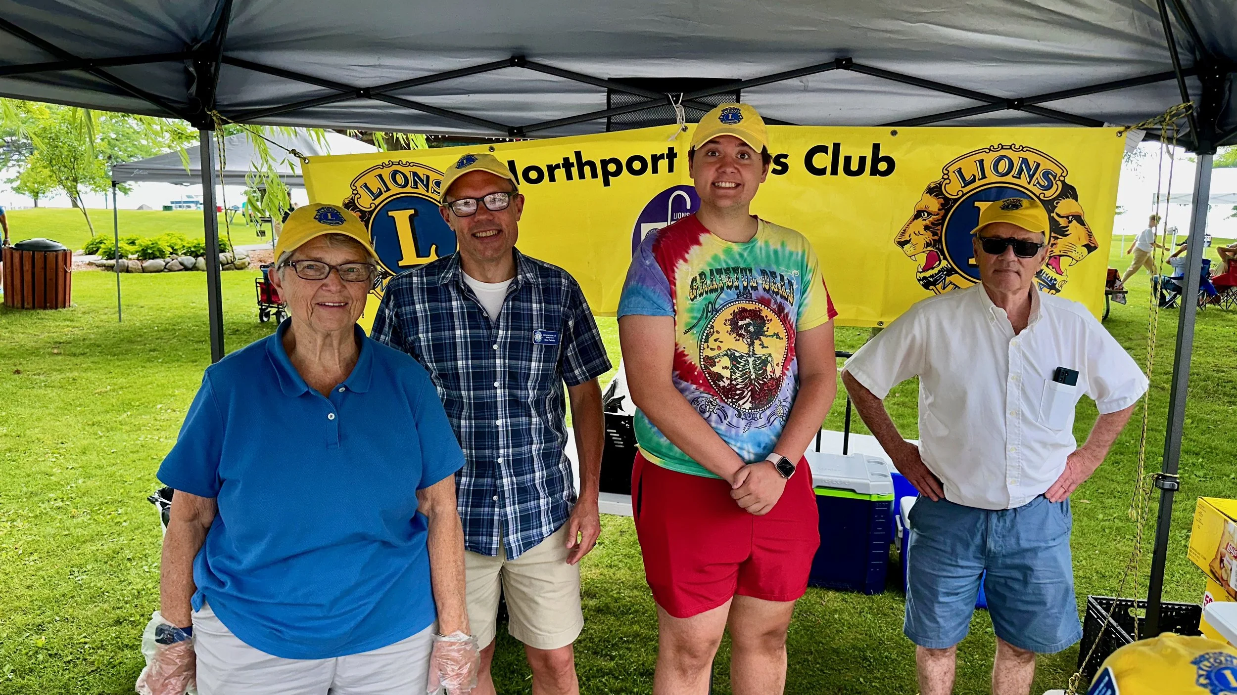 Four smiling people standing in front of a Lions Northport Lions Club banner at an outdoor event. They are under a canopy, with a grassy park and trees in the background.