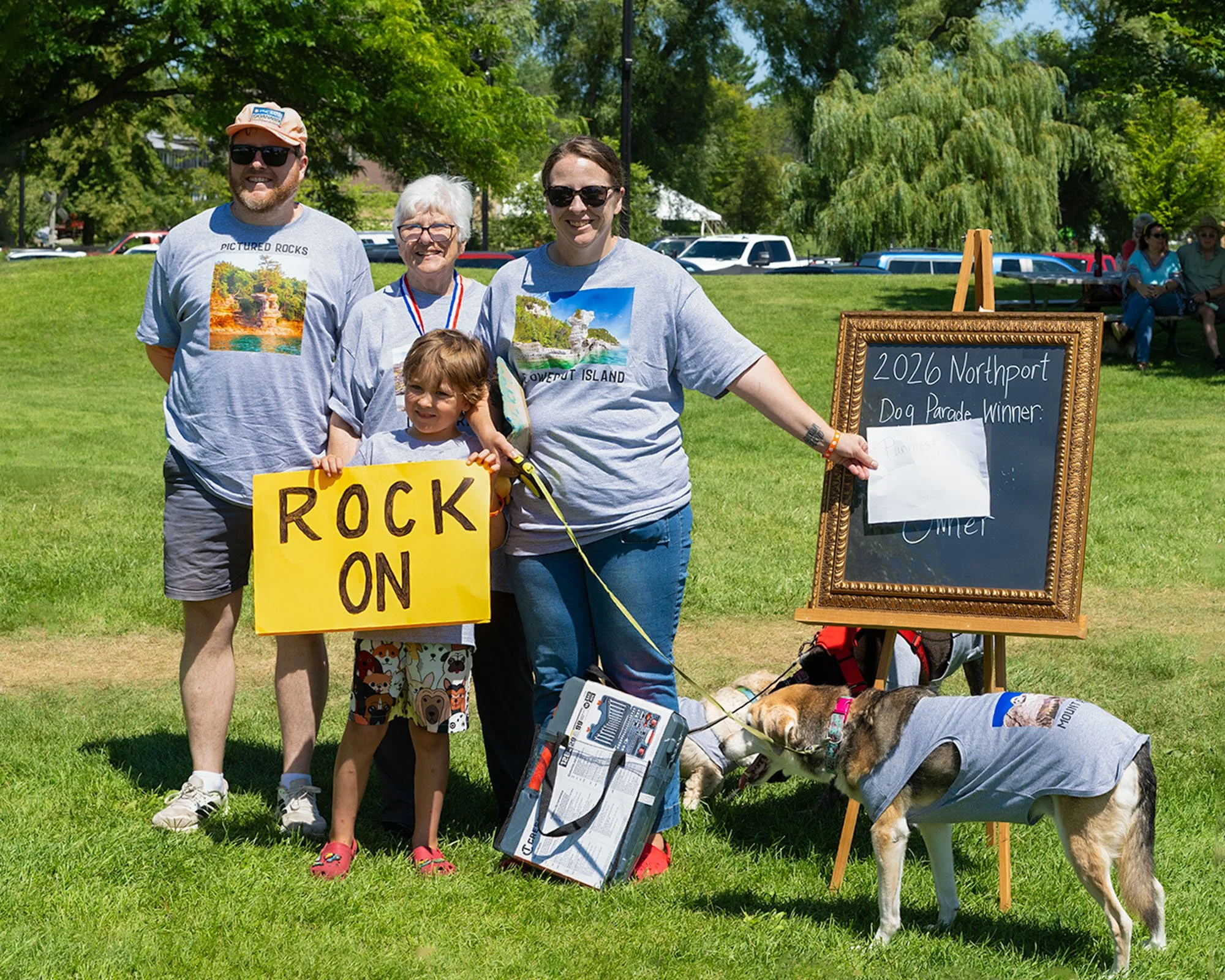 A group of people, including a man, woman, and child, smiling outdoors on a sunny day, holding a yellow sign and standing in front of a chalkboard sign that reads '2026 Northport Dog Parade Winner.' They are accompanied by a beagle dog dressed in a v