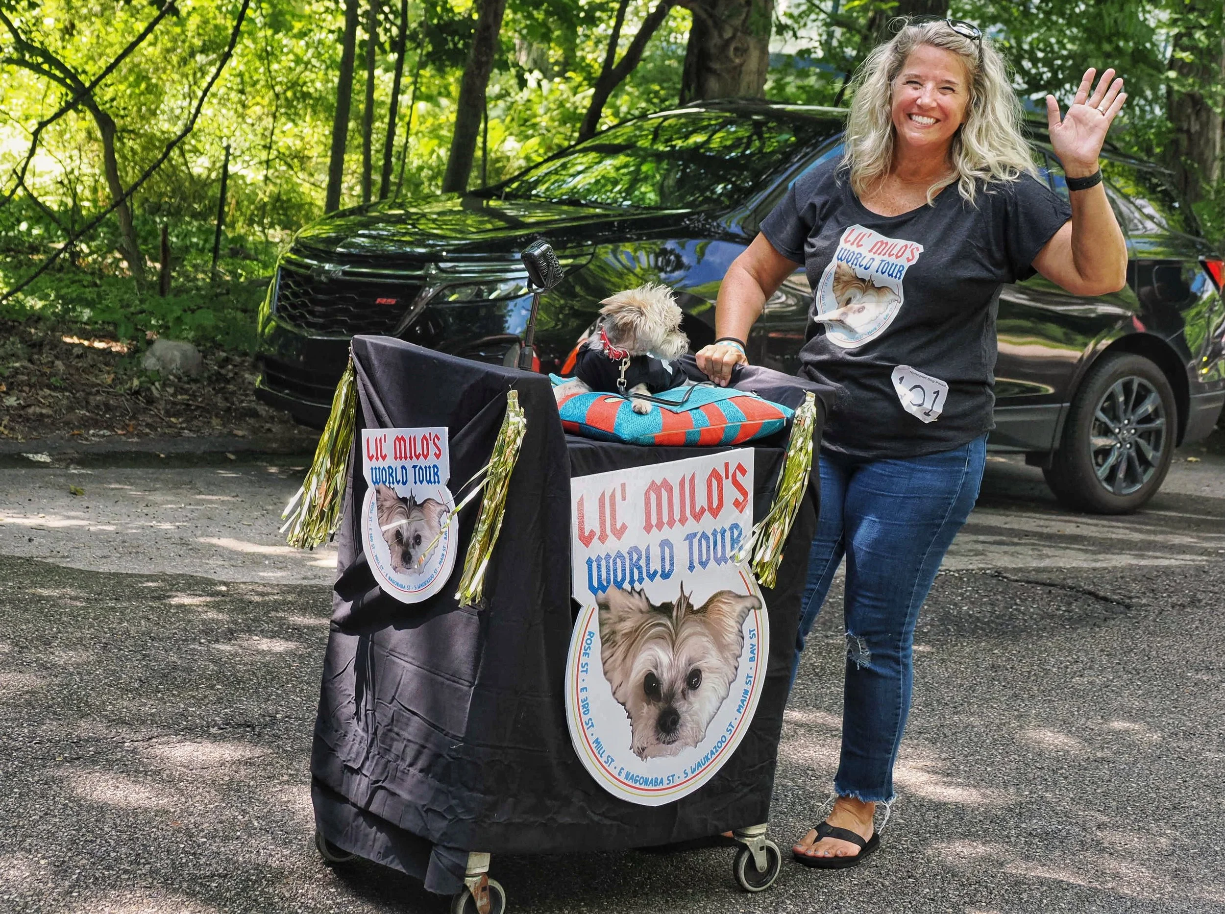 A woman with long curly gray hair, wearing a black T-shirt with 'Lil' Milo's World Tour' printed on it, waving and smiling while standing next to a decorated stroller containing a small dog. The dog is dressed in a black outfit with a red bow and is 