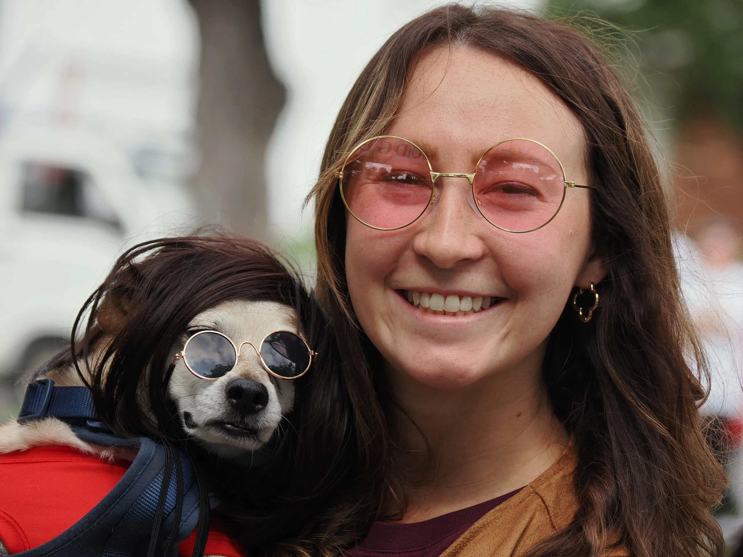 A smiling woman with brown hair wearing round sunglasses and earrings, hugging a small dog with sunglasses, wearing a red harness and being carried in a backpack.