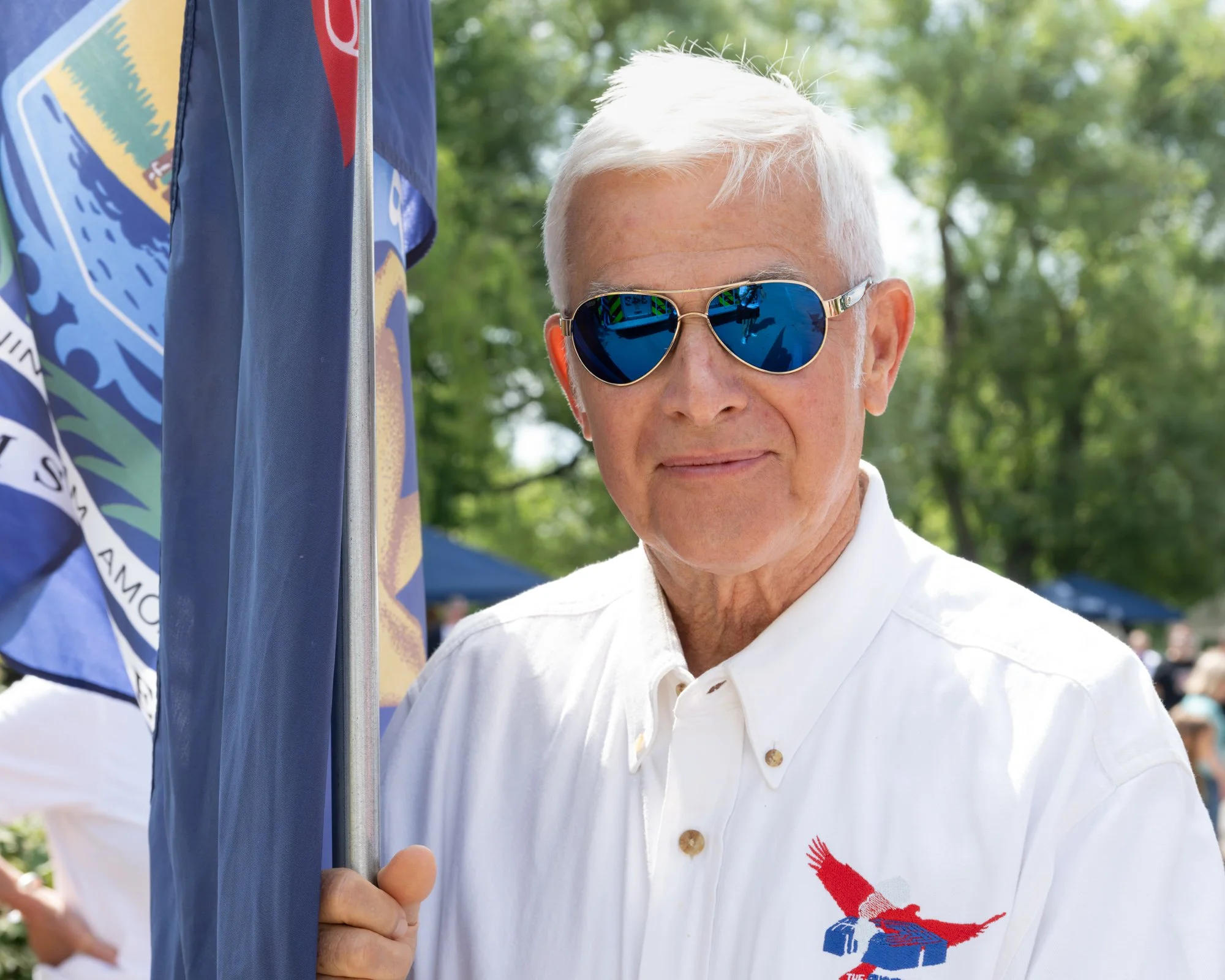 A man with white hair wearing blue reflective sunglasses and a white collared shirt with a red and blue graphic of a lobster or prawn on the shirt, standing outdoors near a flagpole with another flag visible, with green trees and a bright sky in the 