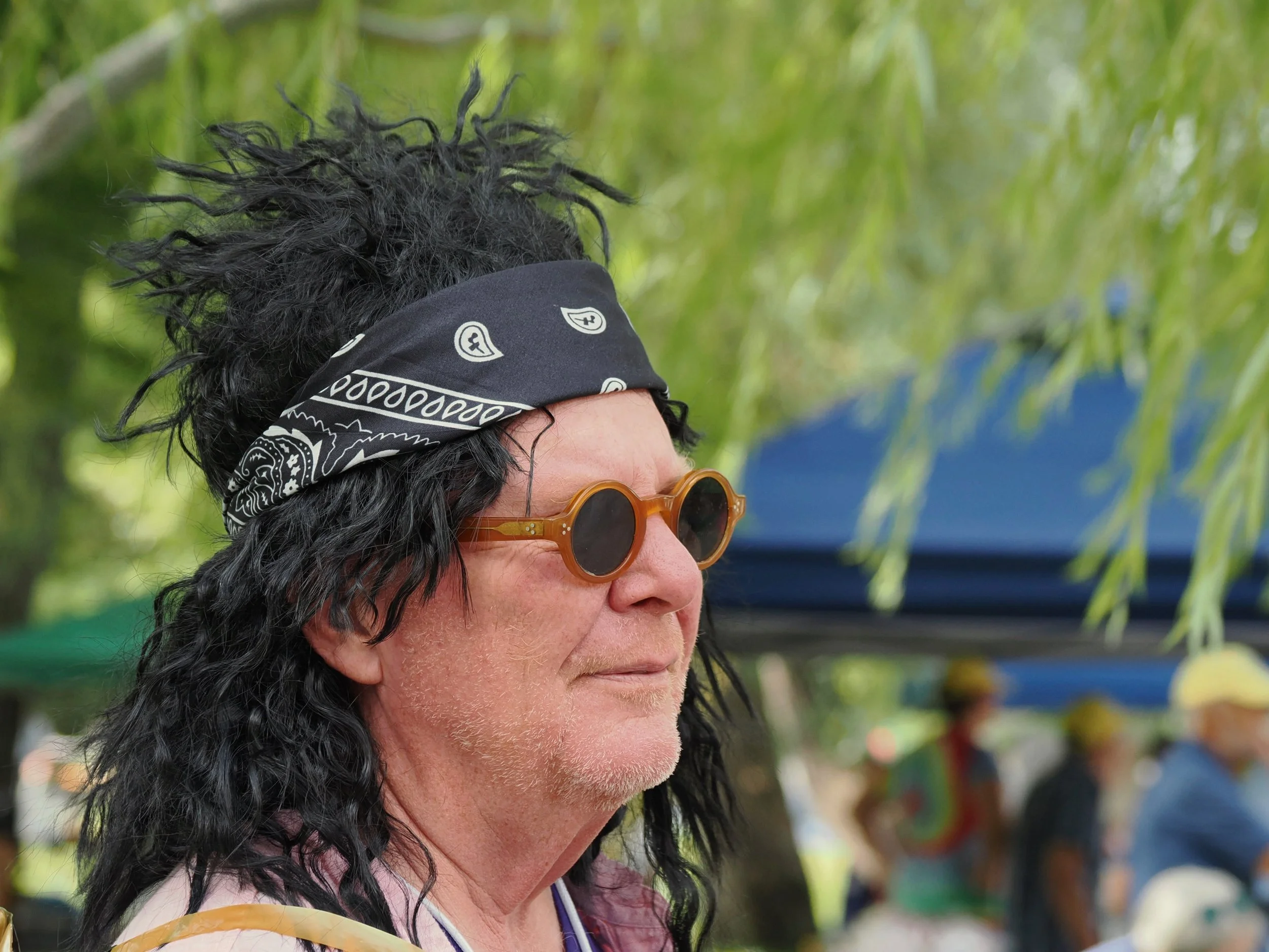 Profile of a middle-aged woman with dark curly hair wearing a black bandana with white patterns, round sunglasses, and a yellow top, outdoors with green trees and blurred background.