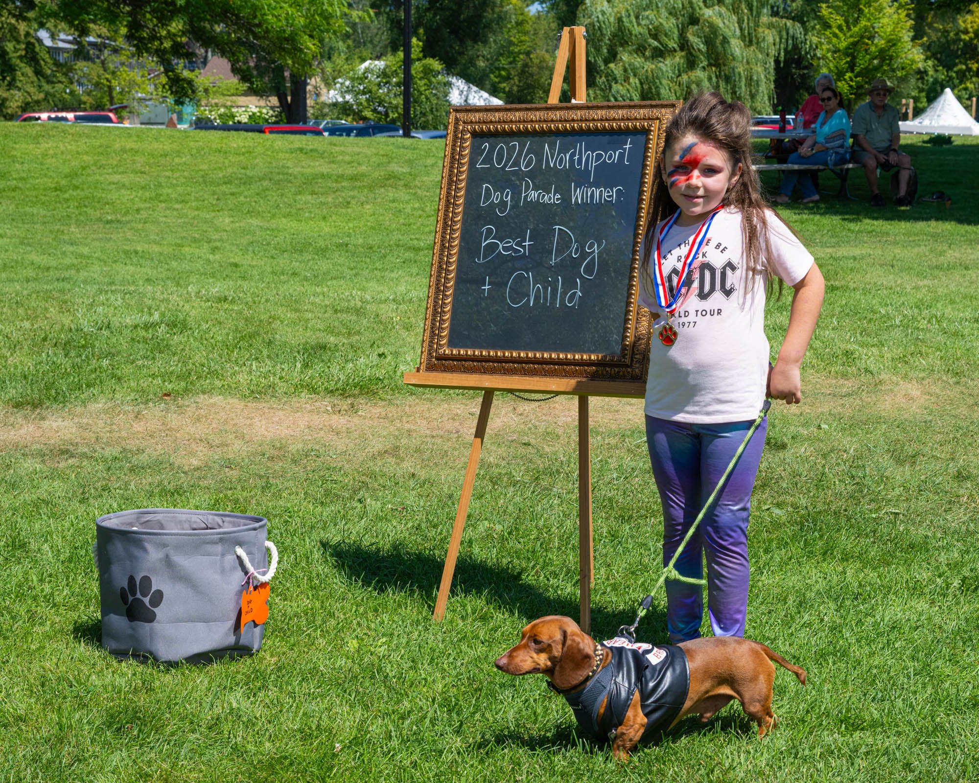 A young girl with face paint standing outdoors on grass with two dogs, one wearing a vest, and a chalkboard sign that reads: '2020 Northport Dog Parade Winner: Best Dog + Child'. In the background, people are sitting on benches under trees, with tent