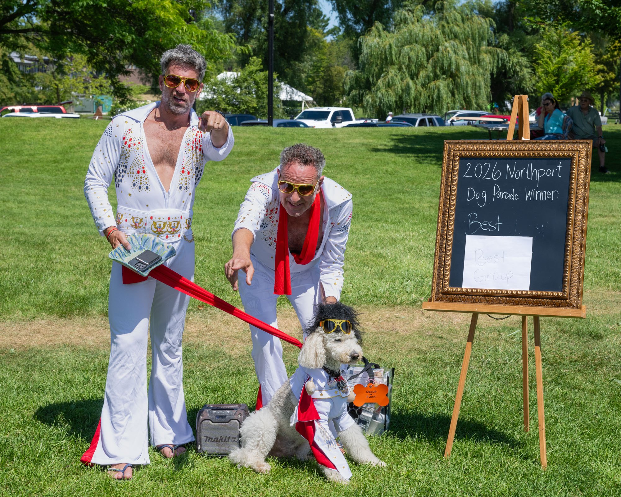Two men dressed in Elvis Presley costumes with sunglasses, one standing and one crouching, with a dog beside them, participating in a dog parade event at a park on a sunny day.