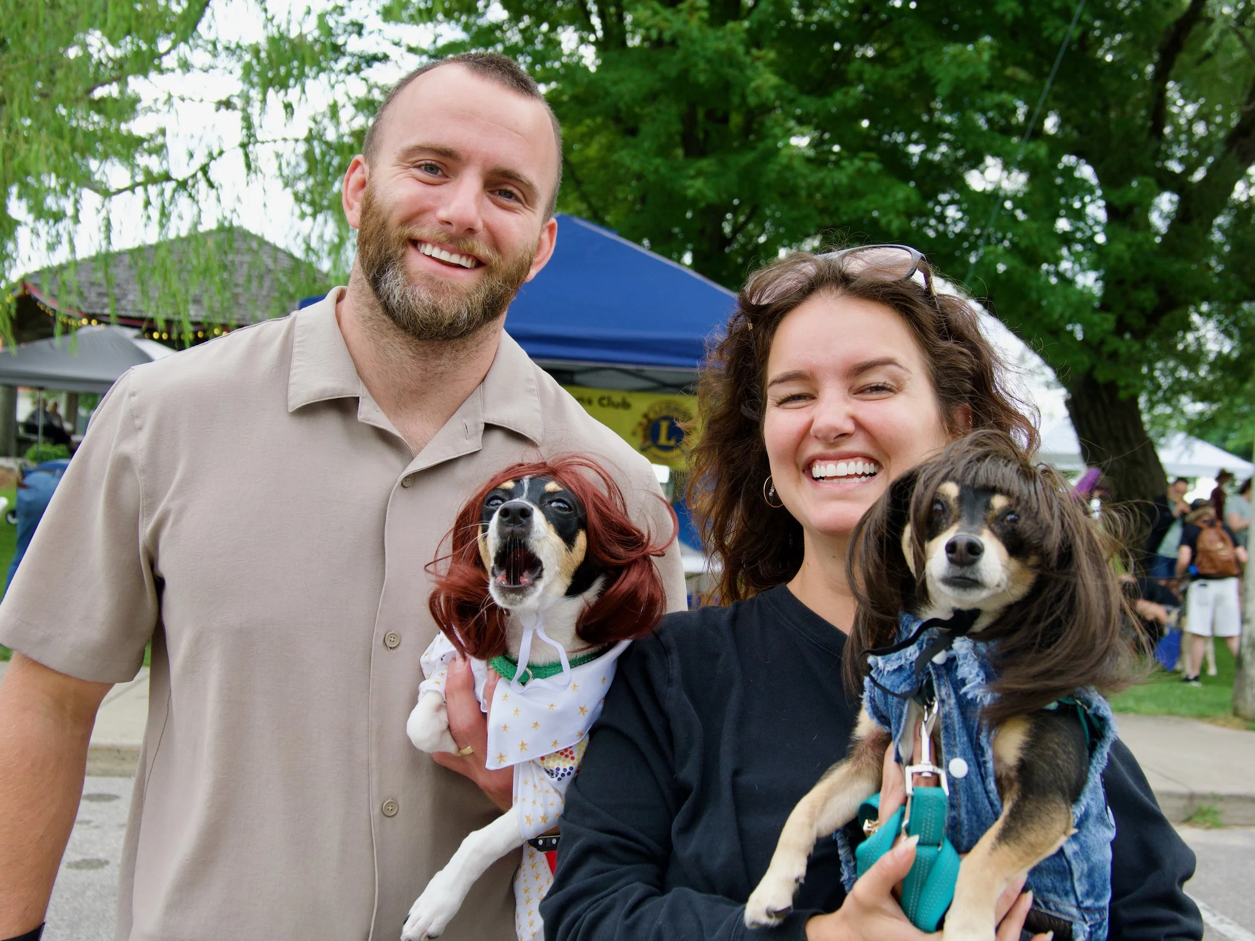 Smiling man and woman holding dogs at an outdoor event with trees and tents in the background.