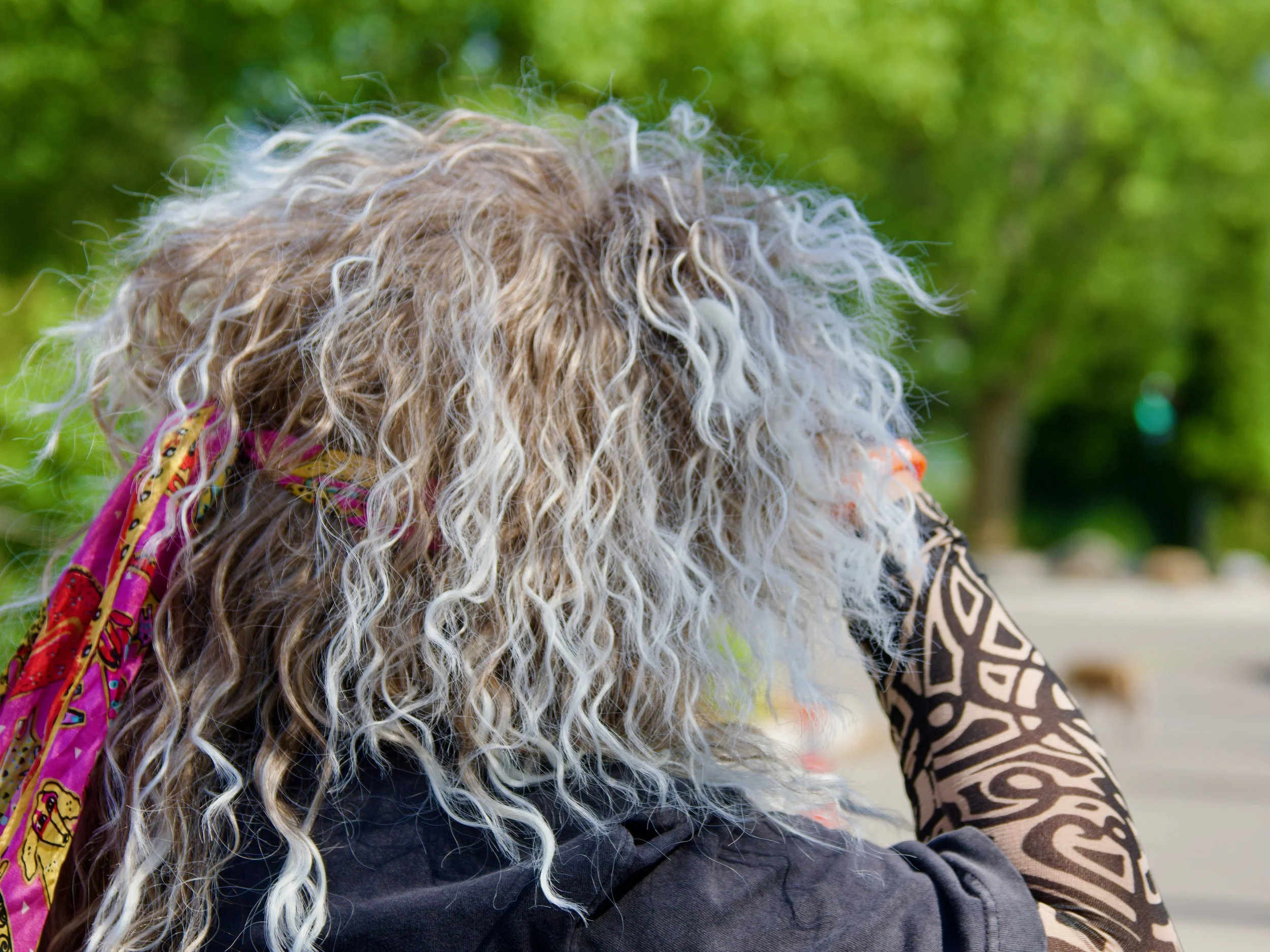 Close-up of a person with long curly blonde hair wearing a colorful scarf and patterned clothing, outdoors with green trees in the background.