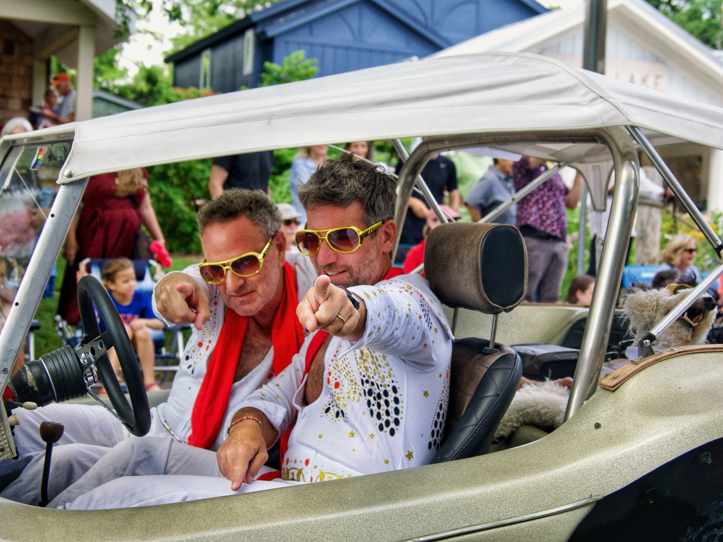 Two men wearing yellow sunglasses sitting in a vintage golf cart at a celebration or parade, with a crowd of people in the background.